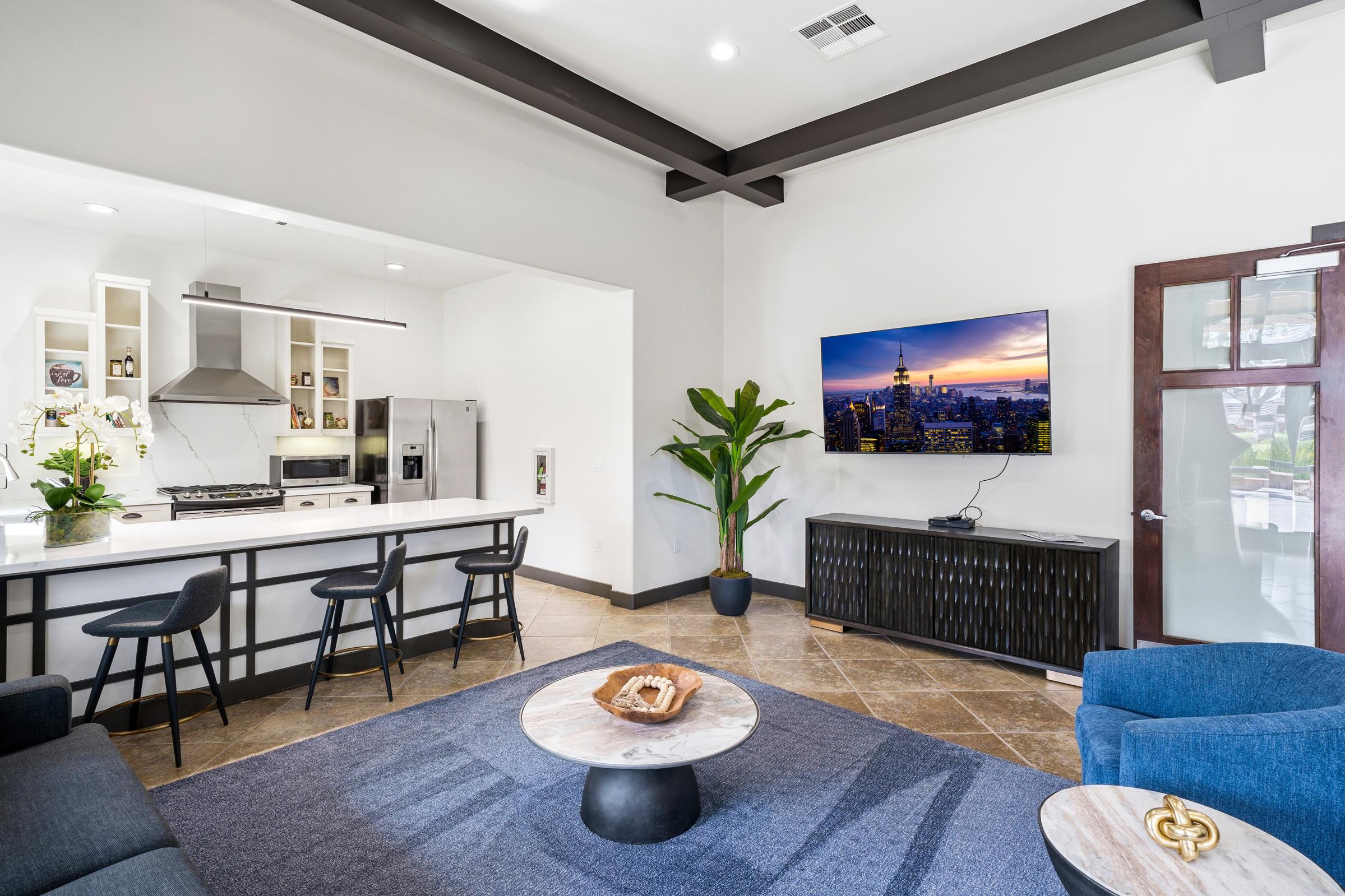 A modern living space featuring a kitchen with a large island and bar seating, a comfortable seating area with a blue chair, and a coffee table. A large wall-mounted TV displays a cityscape. A decorative plant adds a touch of greenery to the contemporary design. Soft lighting and neutral colors create a welcoming atmosphere.