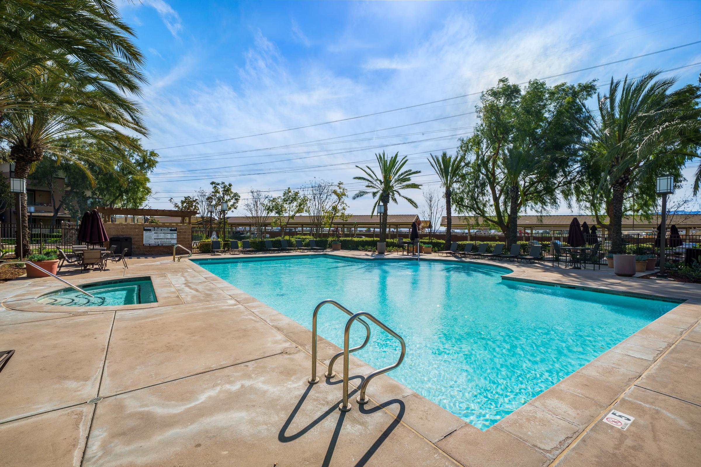 A bright outdoor swimming pool surrounded by palm trees and lush greenery. The pool area features a spacious deck with lounge chairs and shaded umbrellas, set against a clear blue sky. The ambiance suggests a relaxing atmosphere, perfect for leisure and enjoyment.