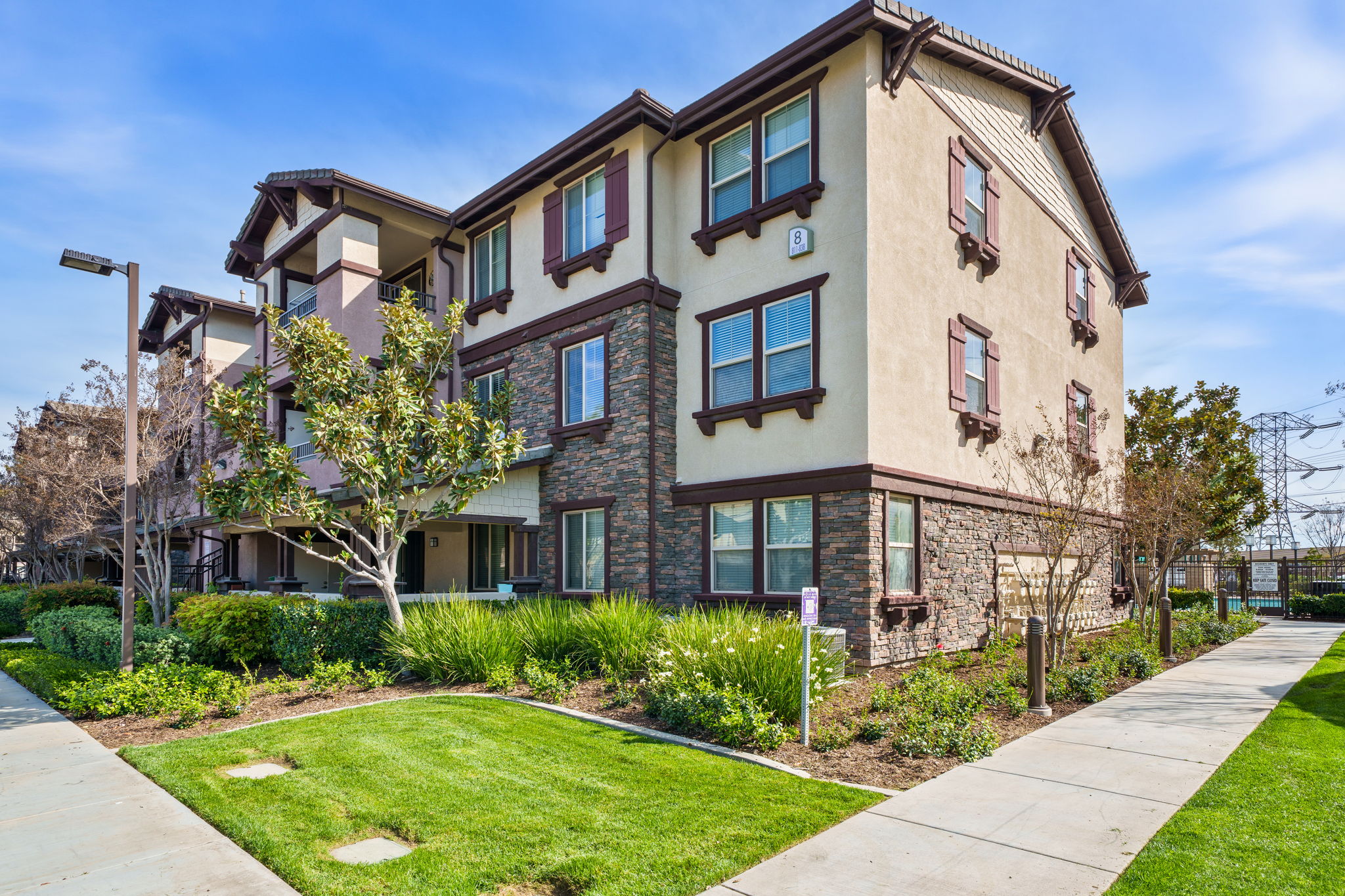 A modern apartment building with a tan and brown exterior. It features multiple windows with decorative trim and a stone accent wall. Surrounding the building are well-maintained green lawns, shrubs, and small trees under a clear blue sky, creating a serene residential atmosphere.