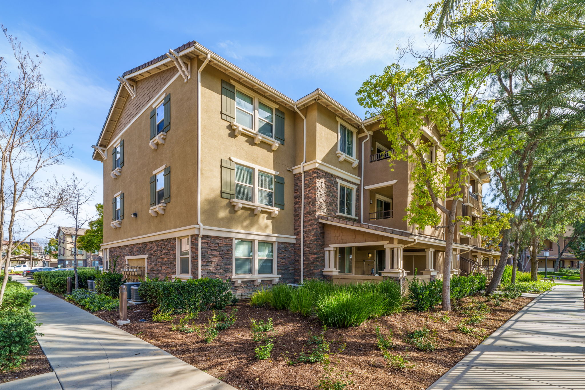 A multi-story residential building featuring a mix of stucco and stone exterior, large windows with green shutters, and landscaped pathways. The setting includes trees and shrubs, contributing to a pleasant outdoor environment on a clear, sunny day.