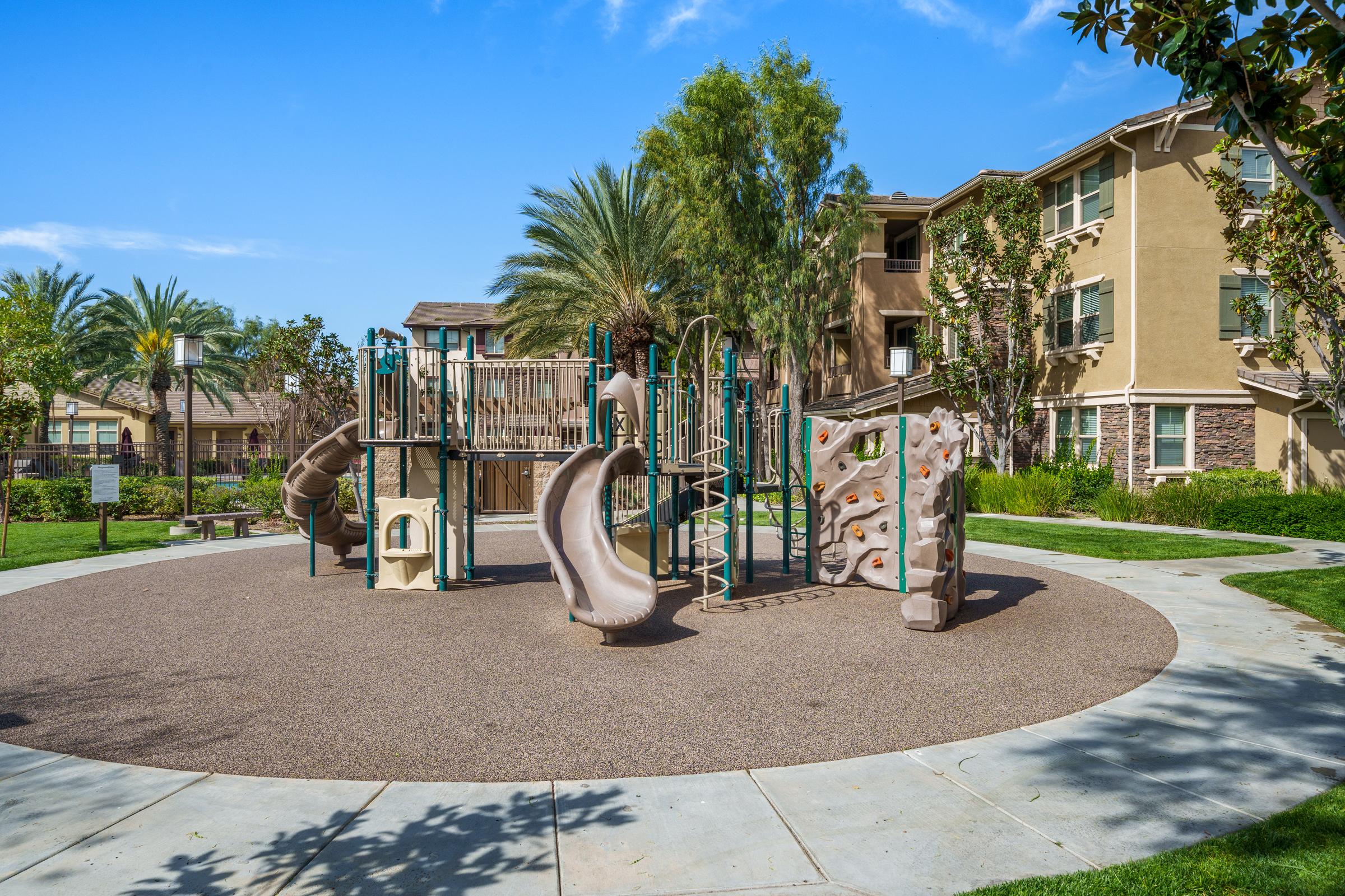 A playground with climbing structures, slides, and safety surfacing, surrounded by green grass and palm trees. Residential buildings can be seen in the background under a clear blue sky.