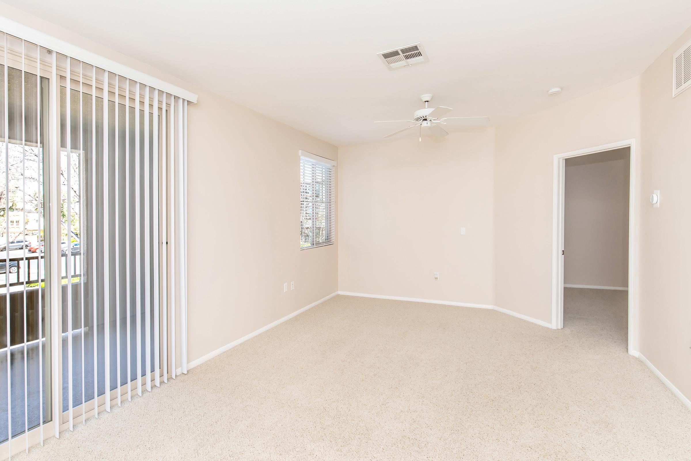 A well-lit, empty living room with beige walls and carpet. Features include a ceiling fan, large windows with vertical blinds, and an open doorway leading to another room. Natural light fills the space, creating a cozy atmosphere. The room is unadorned, offering potential for personal decoration.