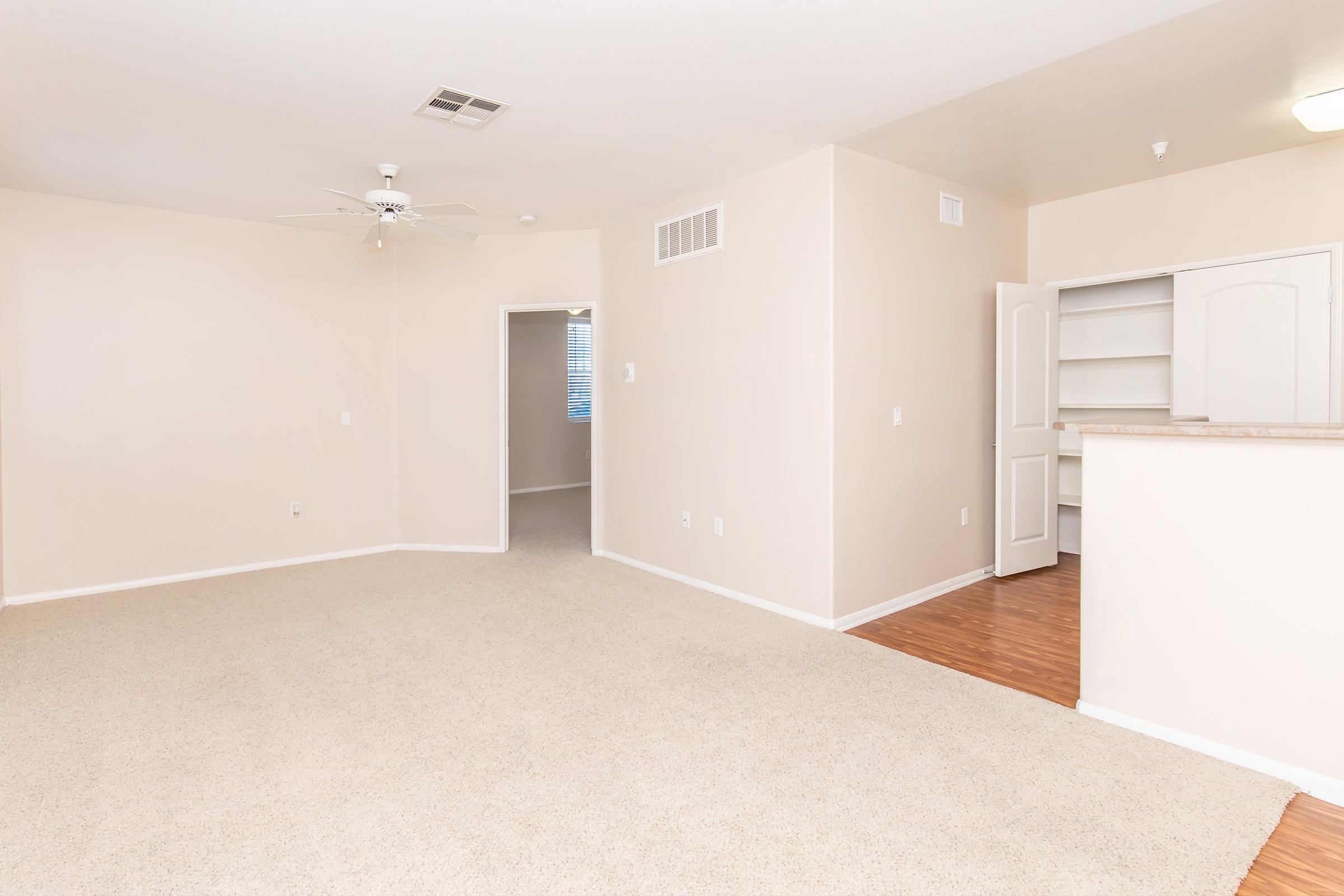 Spacious empty living room with beige walls and light carpet. There is a ceiling fan and an open view of a kitchen area with a bar. A door leads to another room, and a window provides natural light. The overall ambiance is bright and inviting, perfect for a cozy home setting.