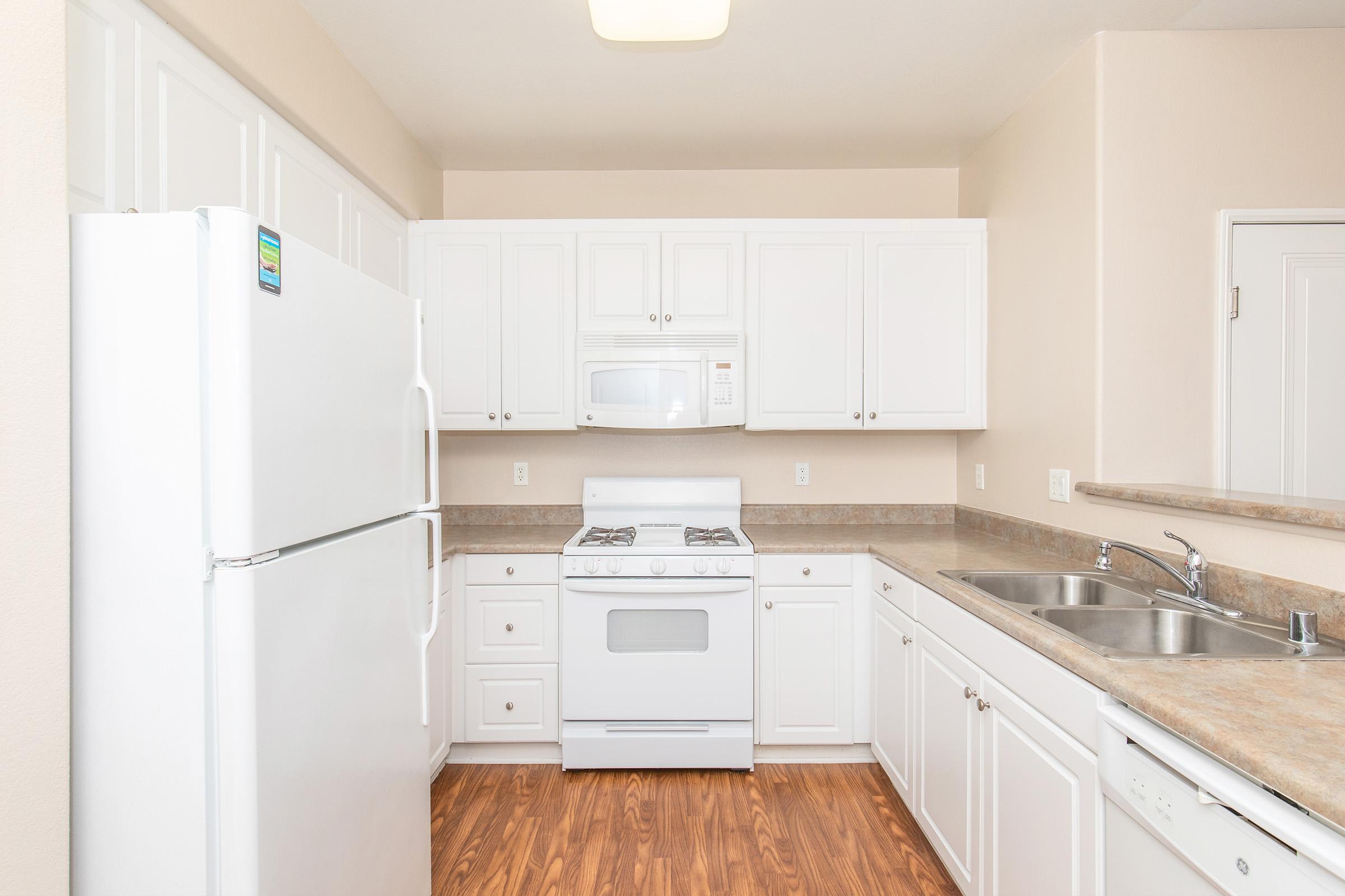 A bright, modern kitchen featuring white cabinetry, a double sink, a gas stove, and a microwave. A large refrigerator is on the left side, with light-colored laminate countertops and hardwood flooring, creating a clean and inviting atmosphere.