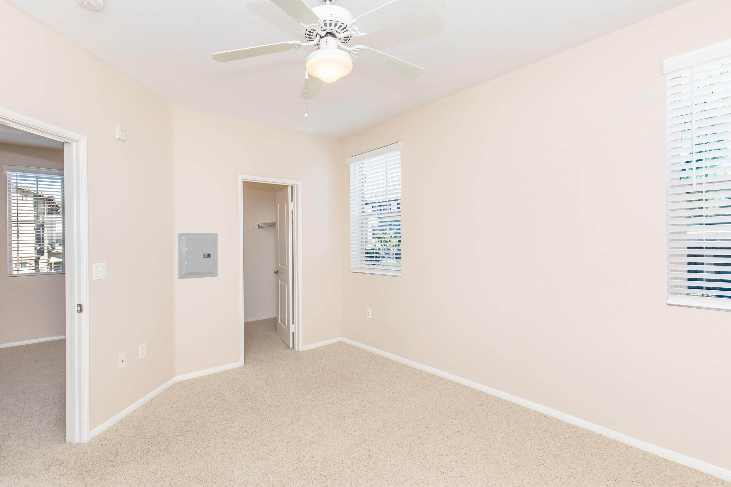 A light-colored room with beige walls and carpet. The room features a ceiling fan, two windows with blinds, and a small closet accessible through an open door. There are two doorways leading to adjacent spaces. The overall atmosphere is bright and tidy.
