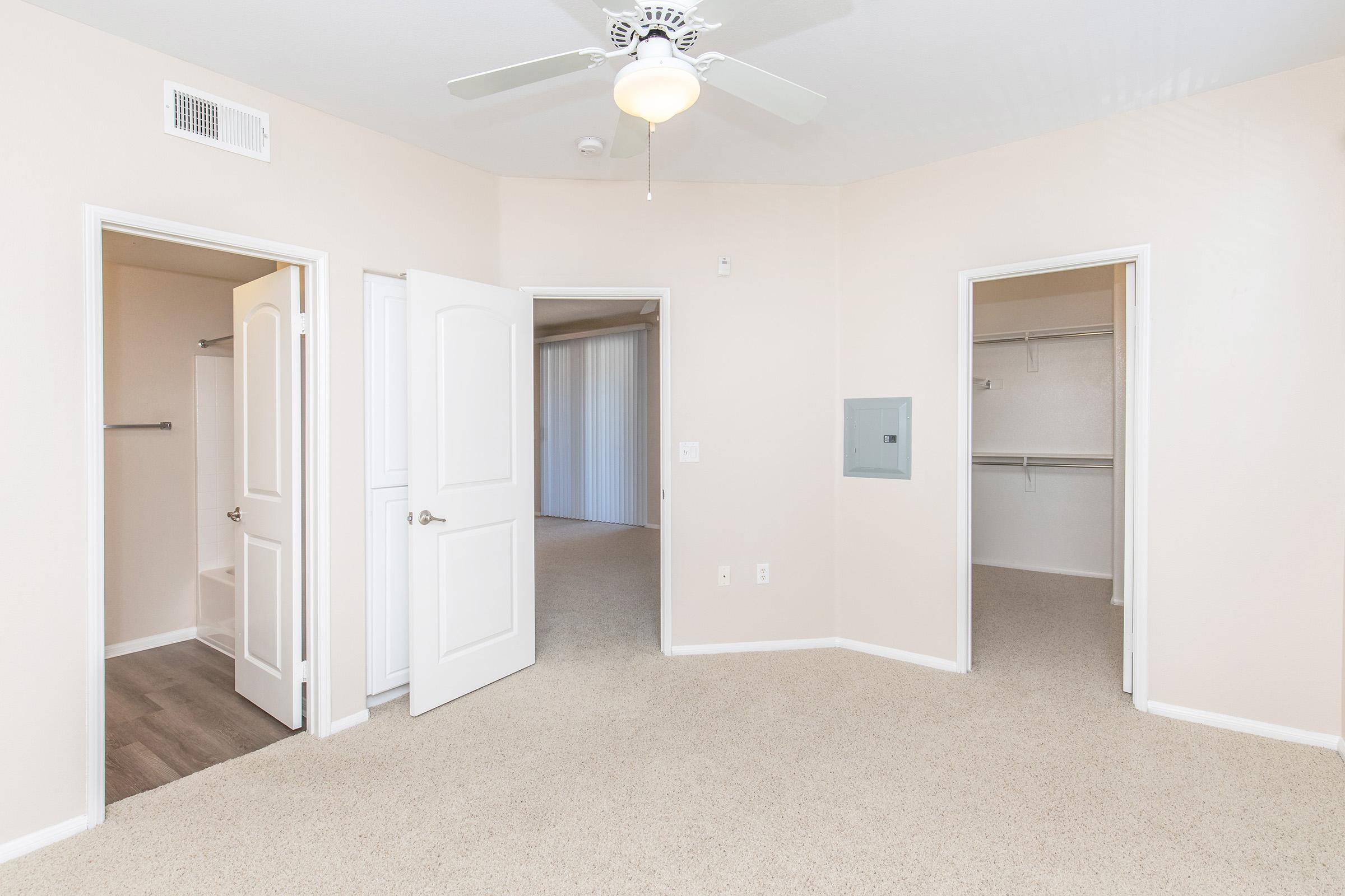 Interior view of a room featuring beige walls and carpet. Two white doors lead to adjacent spaces, one possibly a closet with shelving. A ceiling fan is visible, and there is a light switch on the wall. The overall atmosphere is bright and neutral, suitable for various uses.