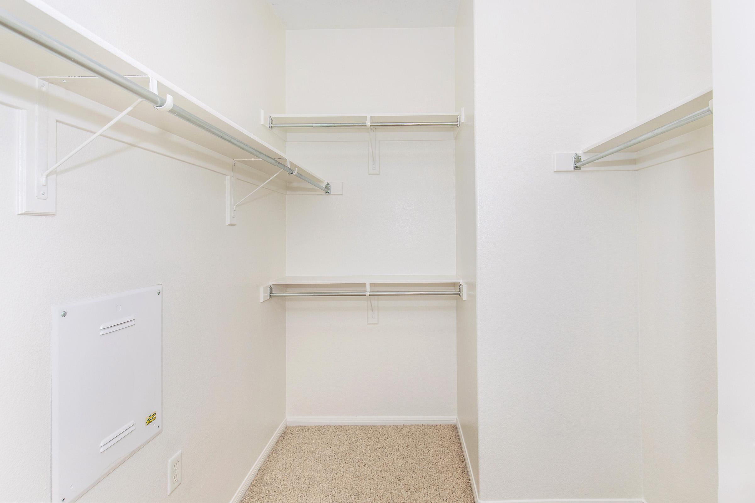 Empty walk-in closet with white walls and light beige carpet. Two hanging rods and a shelf along the back wall, providing ample storage space. The closet has a clean, minimalist look, with no visible items or decorations.