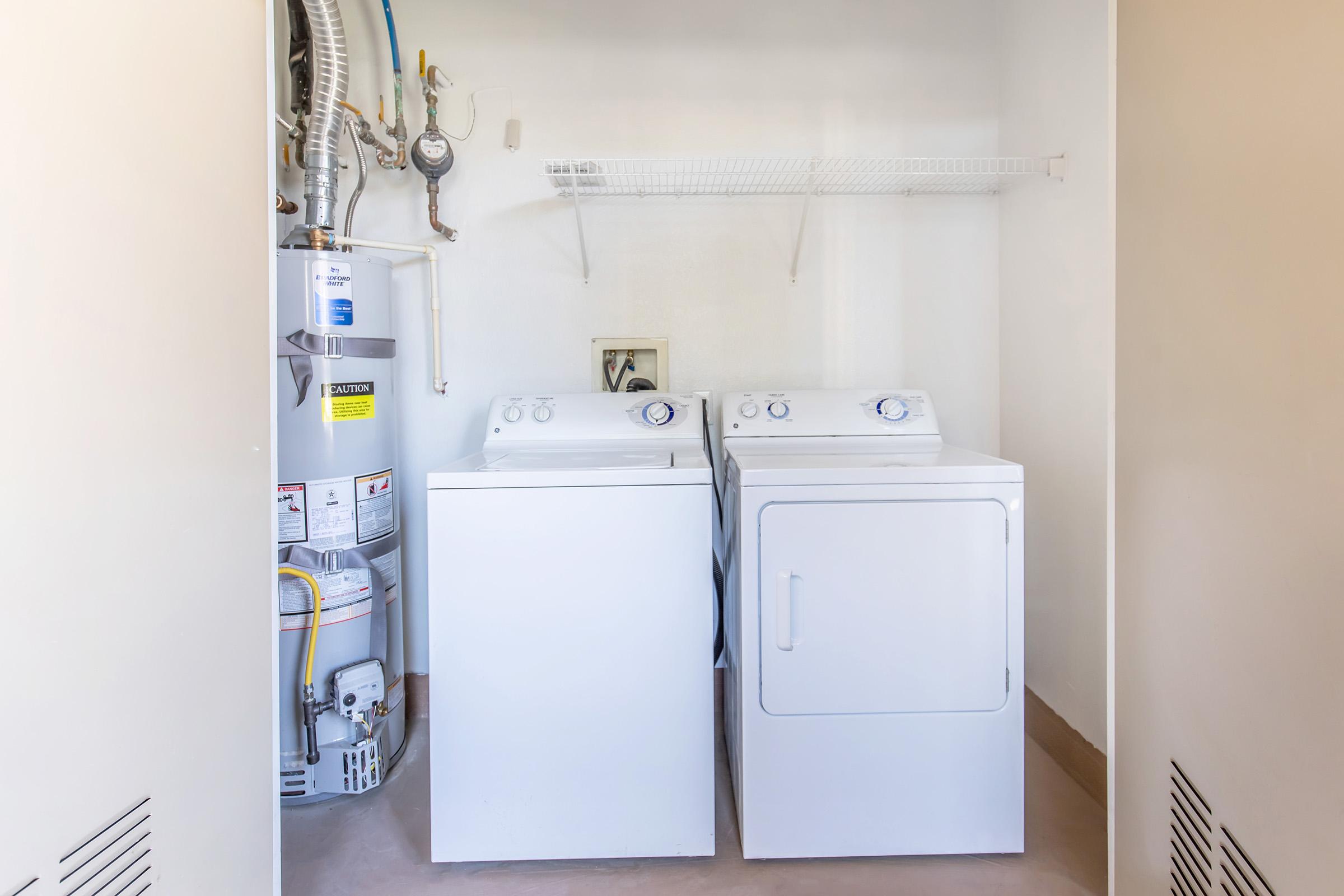 A laundry room featuring a white washer and dryer side by side, with a gas water heater nearby. The space includes a wall shelf above the appliances for storage. The walls are light-colored, enhancing the bright and clean appearance of the room.