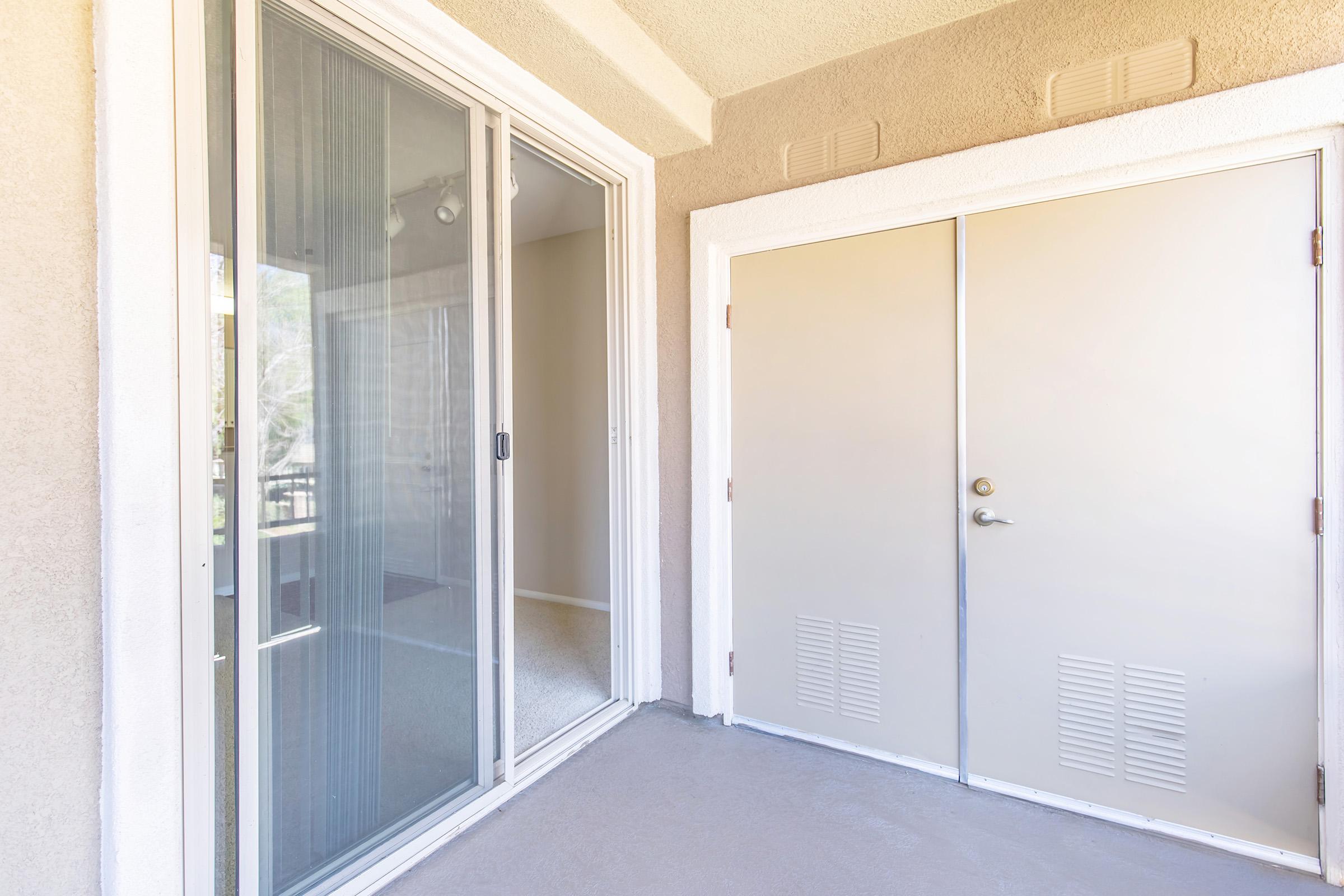 A view of a balcony or patio area featuring a sliding glass door on the left and a double door entrance on the right. The walls are light-colored and there is a concrete floor. Natural light is visible coming from inside the room beyond the glass door.
