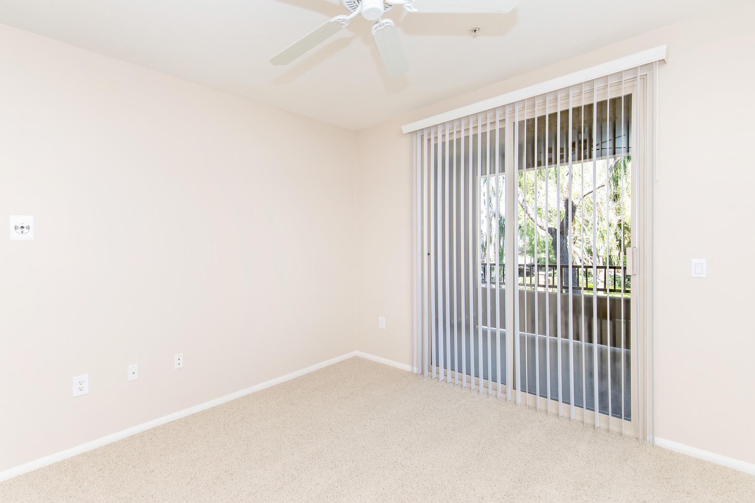 Empty room with light beige walls and cream-colored carpet. A ceiling fan is mounted on the ceiling, and a sliding glass door with vertical blinds leads to a balcony. Natural light filters in from outside, highlighting the spaciousness of the area.