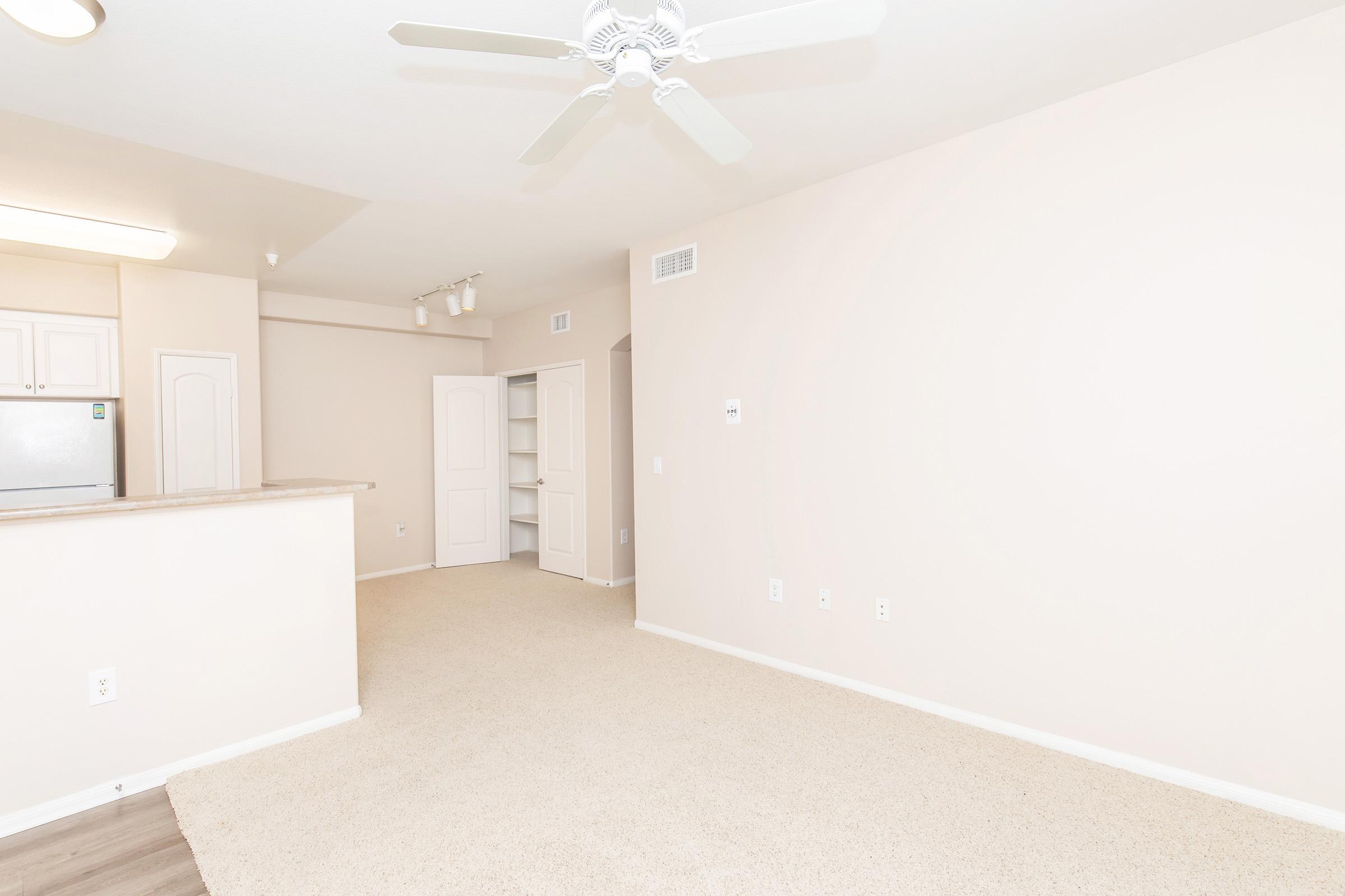 A light-colored living space featuring beige walls and carpet, with a ceiling fan. The area includes a kitchen with a countertop, a door leading to another room, and built-in shelves against one wall. Natural light fills the room, creating a bright and open atmosphere.