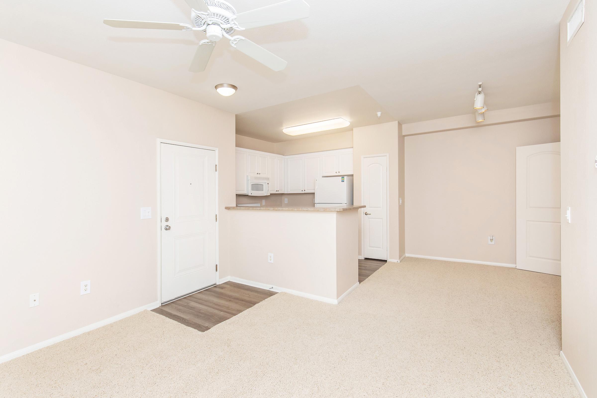 A light-colored living space with beige walls and carpet, featuring a front door on the left, a kitchen area with white cabinets and a small counter, and an open layout leading to an adjacent area. A ceiling fan and overhead lighting add brightness to the room.