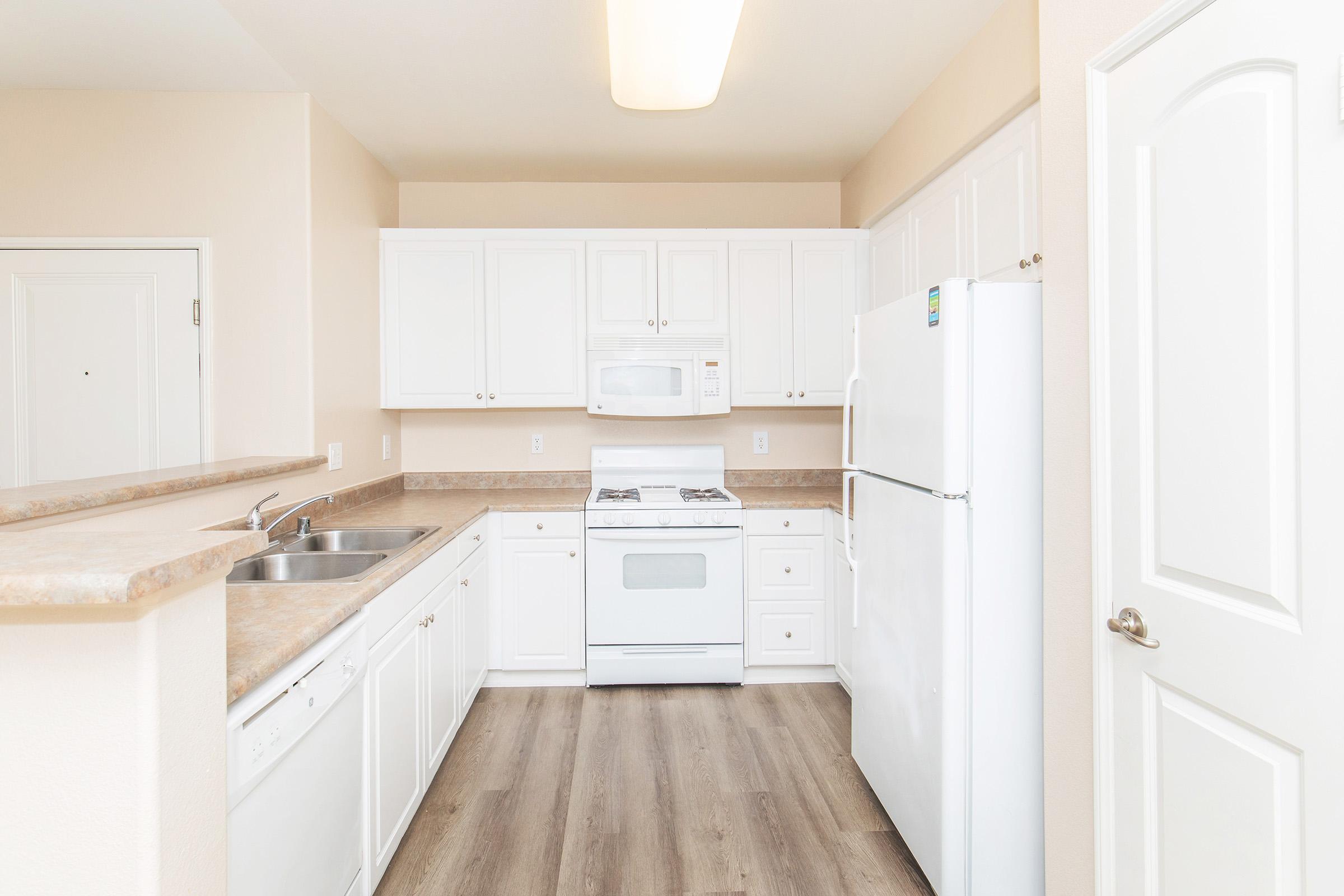 A modern kitchen featuring white cabinetry, a gas stove, a double sink, and a refrigerator. The countertops are light-colored, and the flooring is a wood-like laminate. Soft lighting illuminates the space, creating a clean and inviting atmosphere. The walls are painted in a neutral tone.