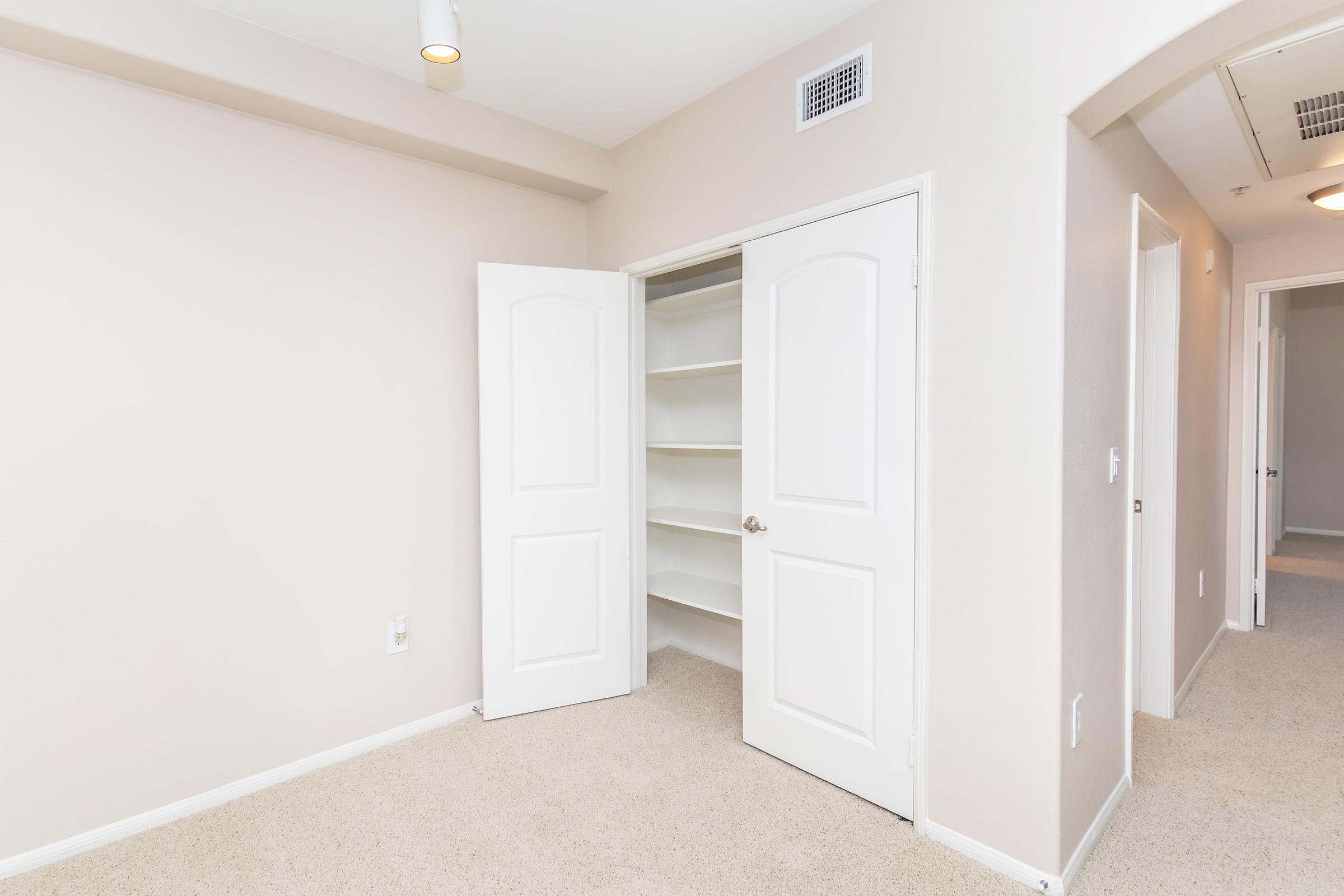 A well-lit room featuring a closet with open double doors. Inside the closet are several shelves. The walls are painted a light color, and the carpeted floor is neutral. There are additional doorways leading to other areas of the space, creating a spacious and organized appearance.