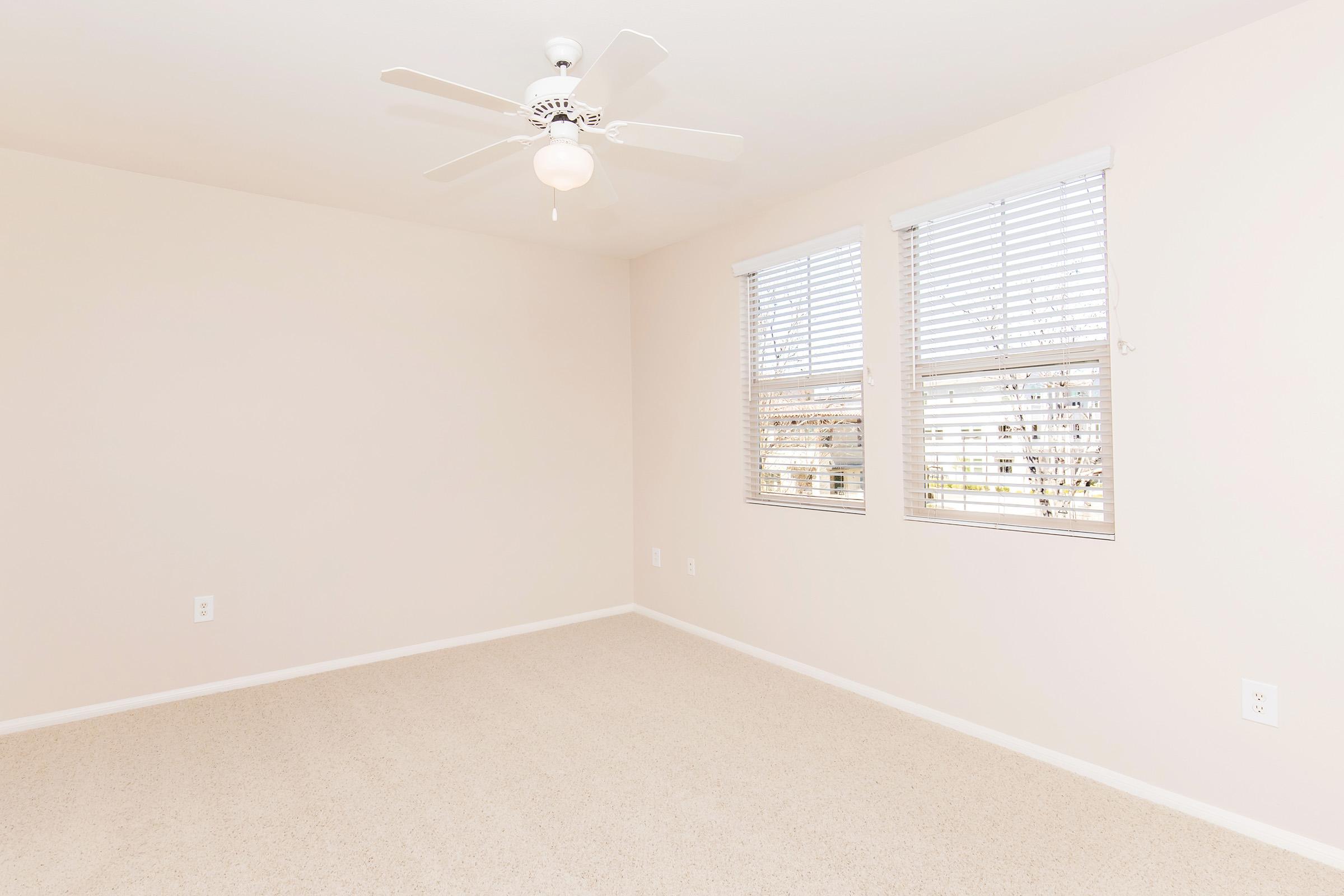 Empty room with beige walls and light-colored carpet. Two windows with white blinds allow natural light to enter. A ceiling fan with a light is centered in the room, enhancing the neutral and airy atmosphere. No furniture is present, creating a spacious and open feel.