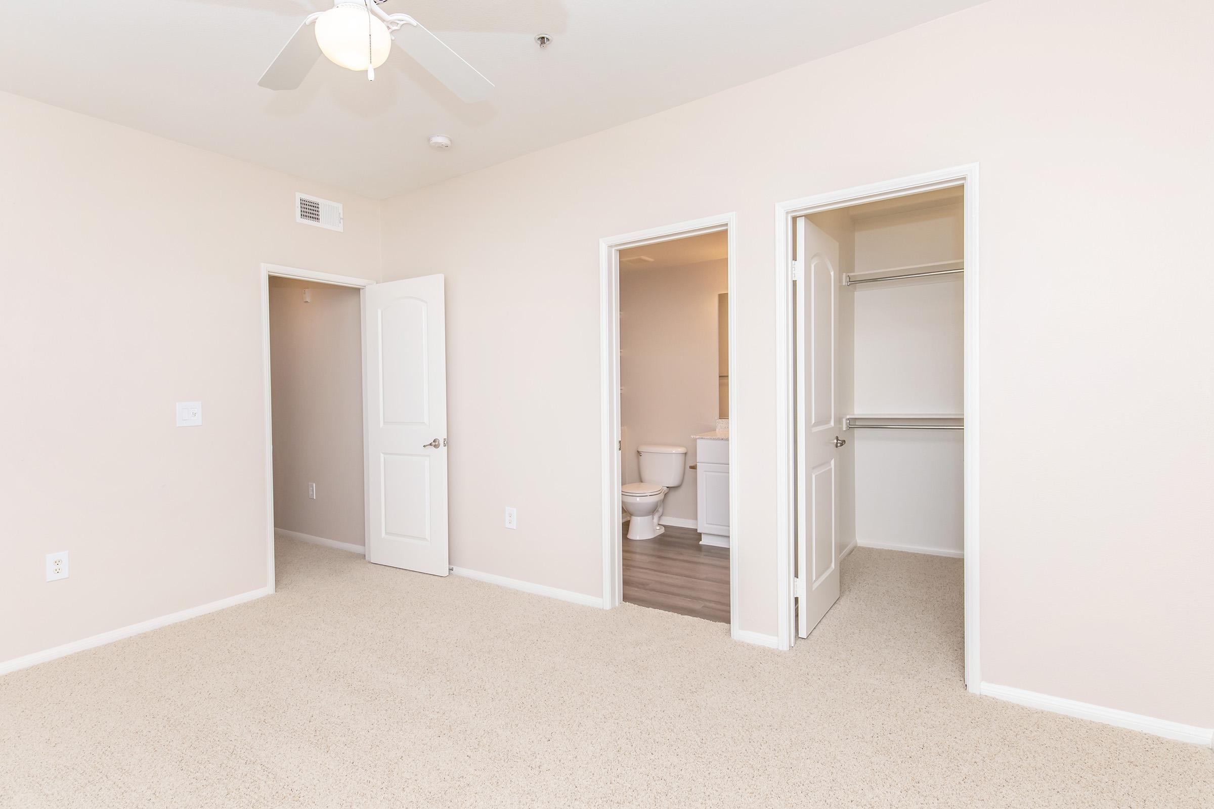 A spacious room with beige walls and carpet. Two doors lead to a bathroom and a closet. Ceiling fan mounted, with natural light illuminating the space.