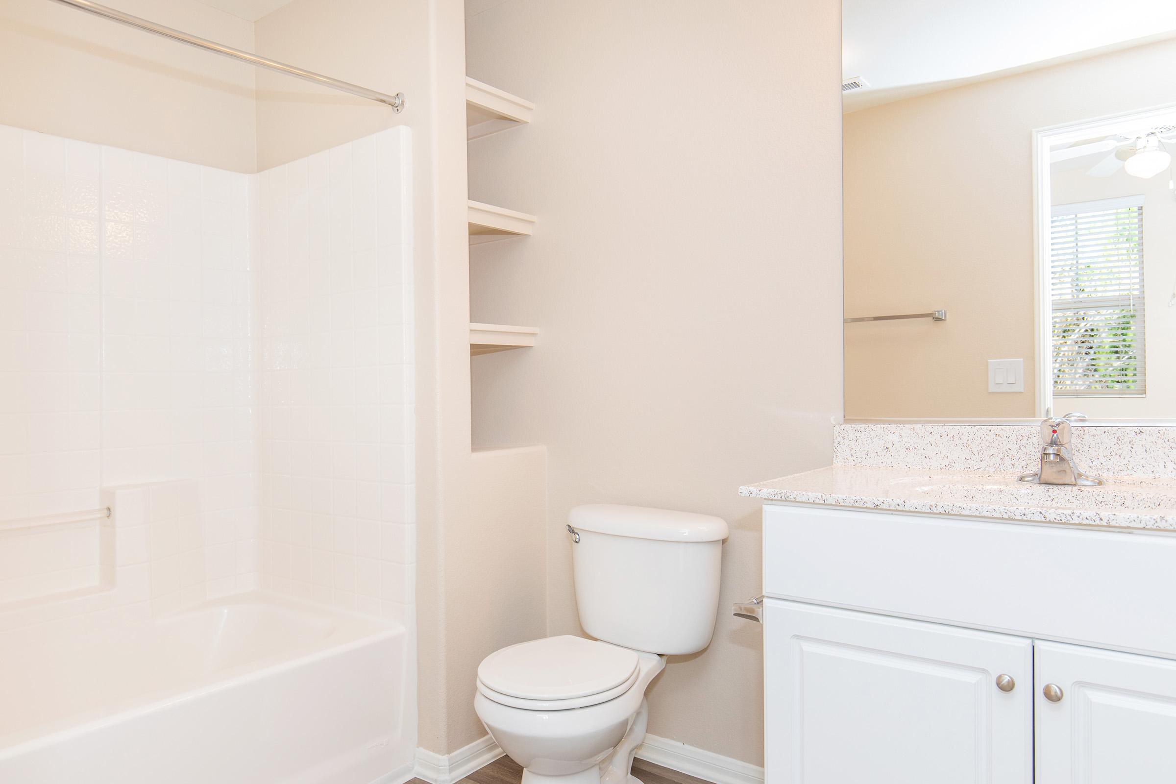 A clean and modern bathroom featuring a white bathtub with a shower, a white toilet, and a sink with a granite countertop. The walls are painted in a soft beige color, and there are built-in shelves next to the bathtub. A mirror is reflected in the light from a nearby window.