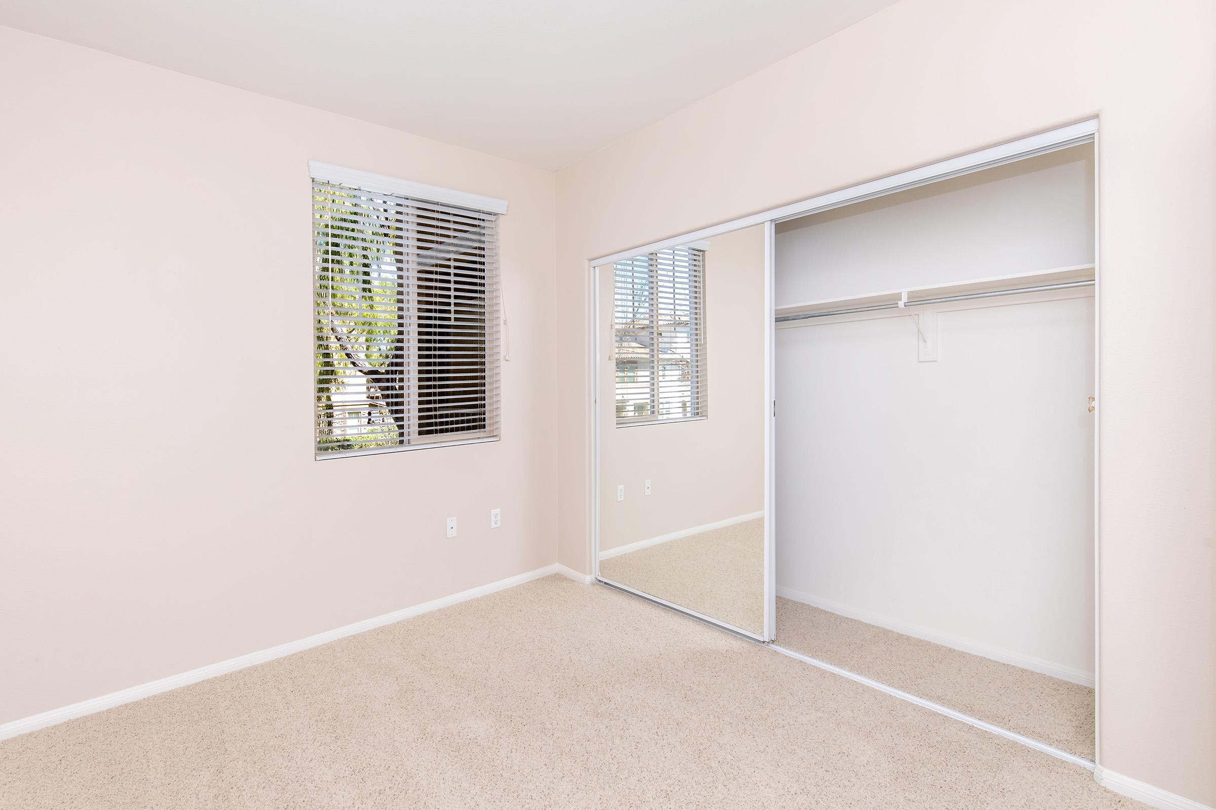 Empty room with light beige walls and beige carpet. A window with blinds on the left allows natural light in. To the right, a mirrored closet with sliding doors reflects the room, and there’s an empty closet space inside. The overall atmosphere is clean and inviting, suitable for personal use or decoration.