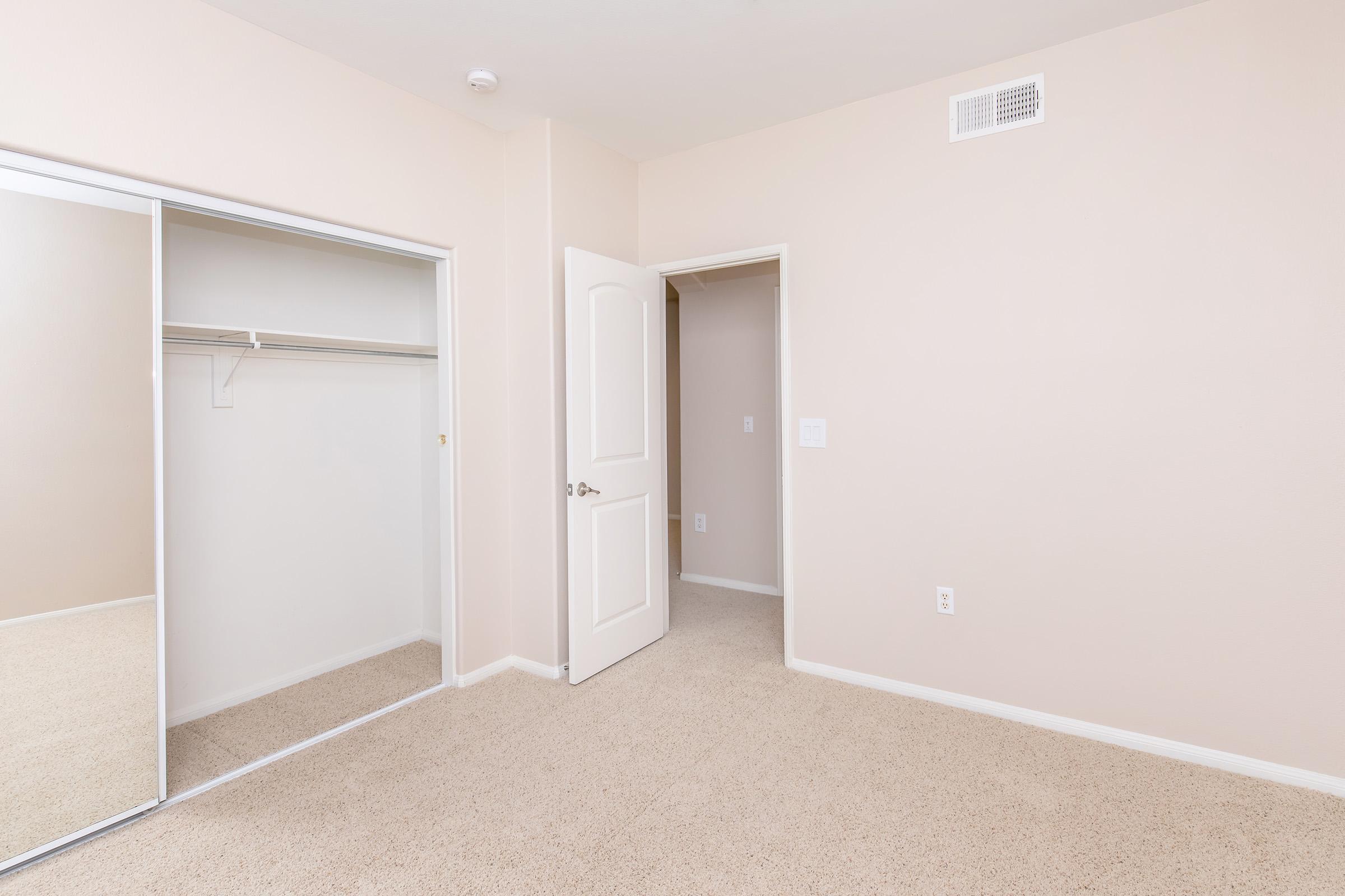 An empty room featuring light beige walls and carpet. A closet with mirrored sliding doors is visible on the left, while an open door leads to another room on the right. The space is brightly lit and appears clean and uncluttered.