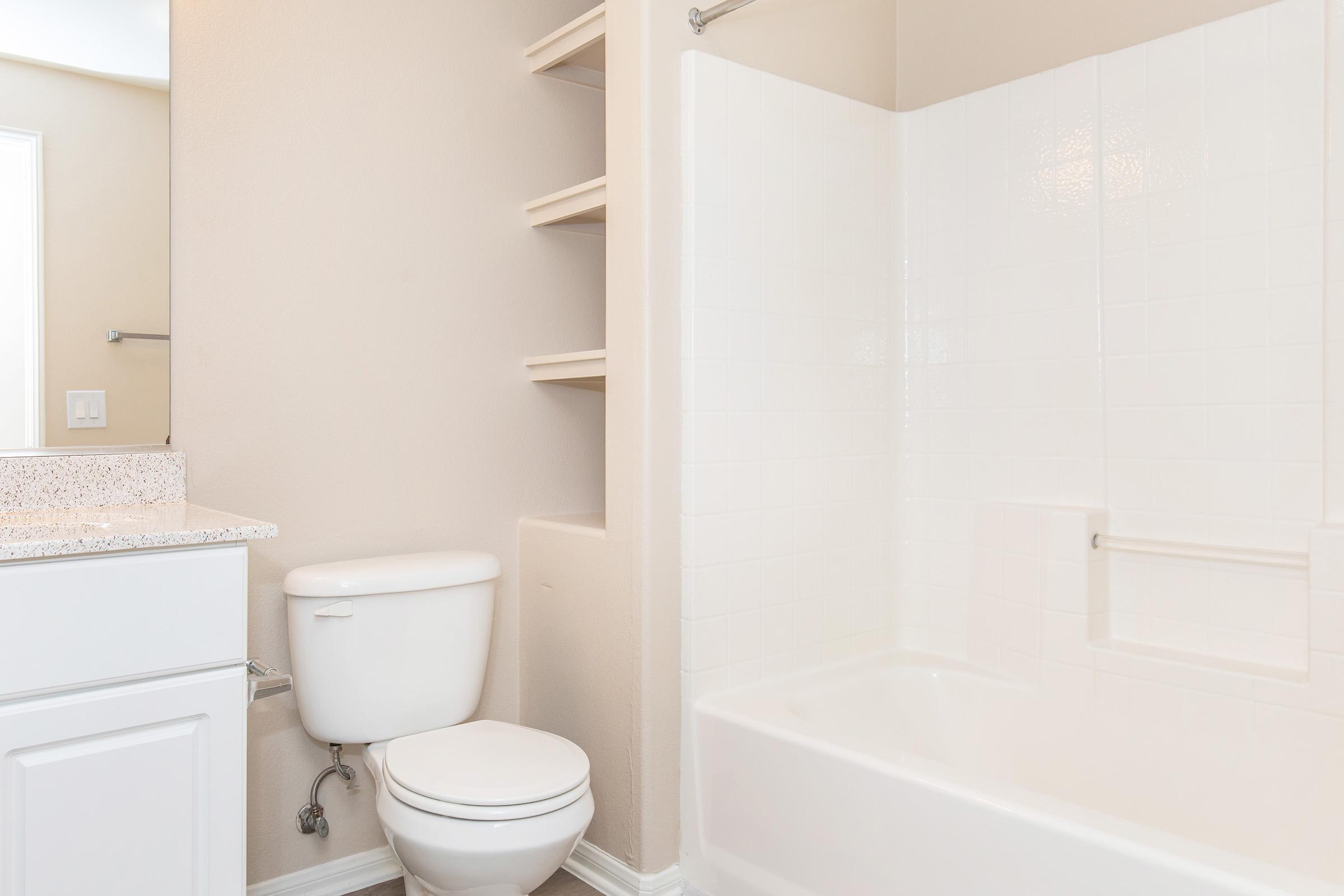 A clean and modern bathroom featuring a white bathtub with a shower, a toilet, a sink with a granite countertop, and open shelving on the wall. The walls are painted a light neutral color, and there is a mirror above the sink.
