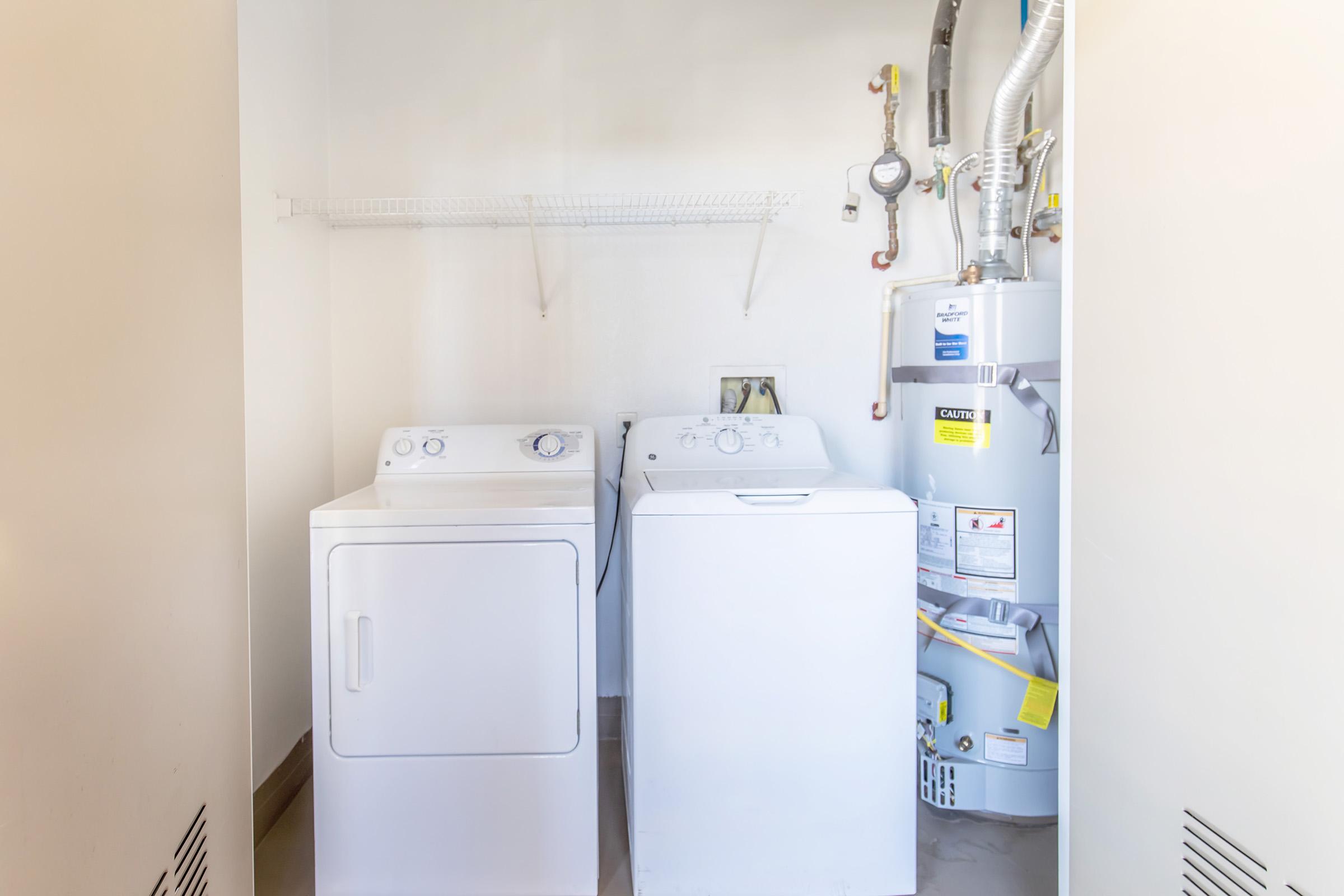 A clean laundry room featuring a stacked washing machine and dryer side by side, with a water heater in the corner. The walls are plain and light-colored, and there is a wire shelf above the appliances. The floor is smooth and uncluttered, creating a tidy, functional space.