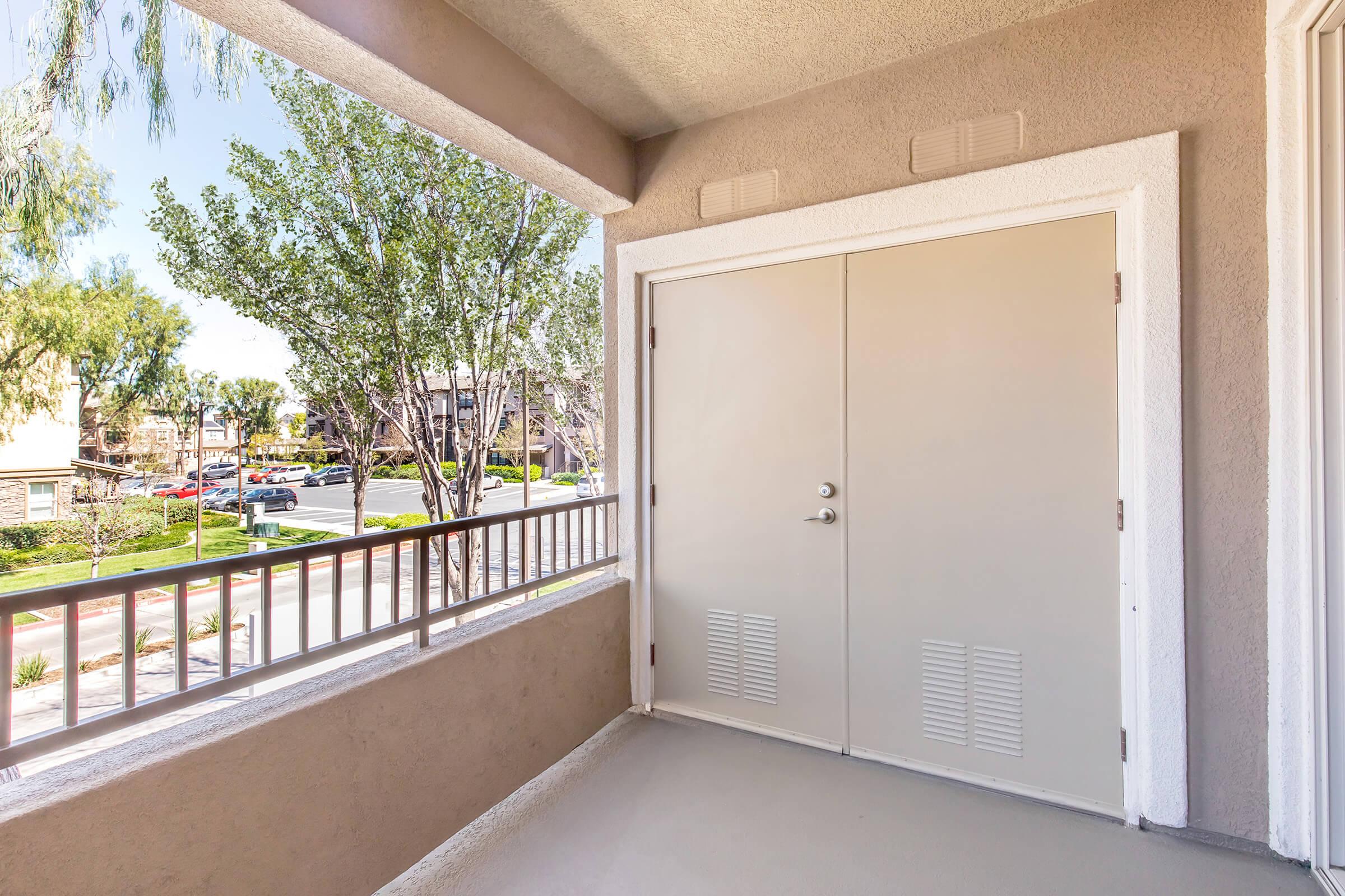 A view of a balcony with beige walls and a closed door. The balcony features a railing and trees are visible in the background, along with a street lined with parked cars. Bright blue sky and sunlight illuminate the scene, creating a welcoming outdoor space.