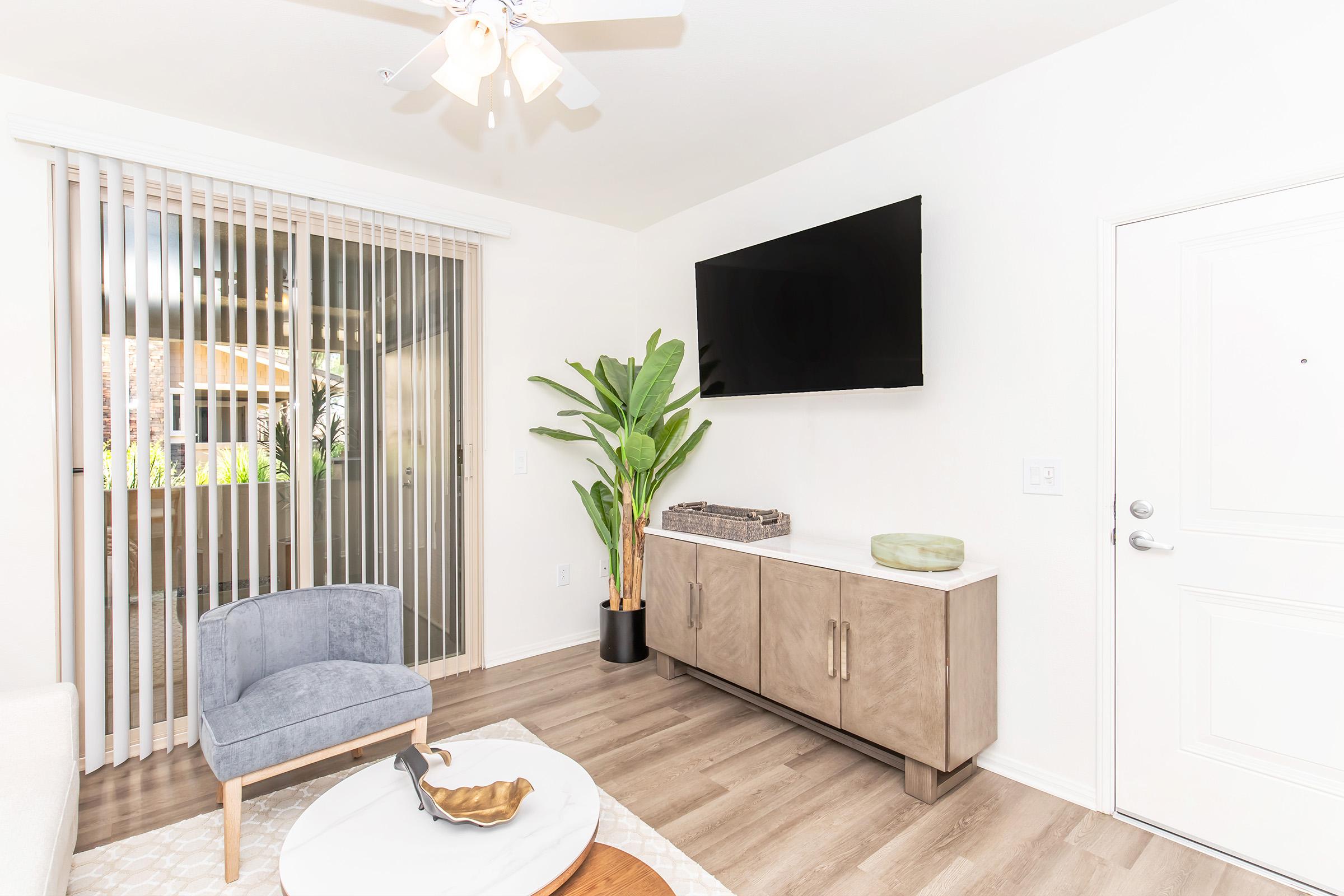 A modern living room featuring a light-colored sofa, a gray armchair, a round coffee table, and a stylish TV mounted on the wall. Large sliding glass doors with vertical blinds allow natural light, and a decorative plant adds a touch of greenery to the space. The floor is made of light wood.