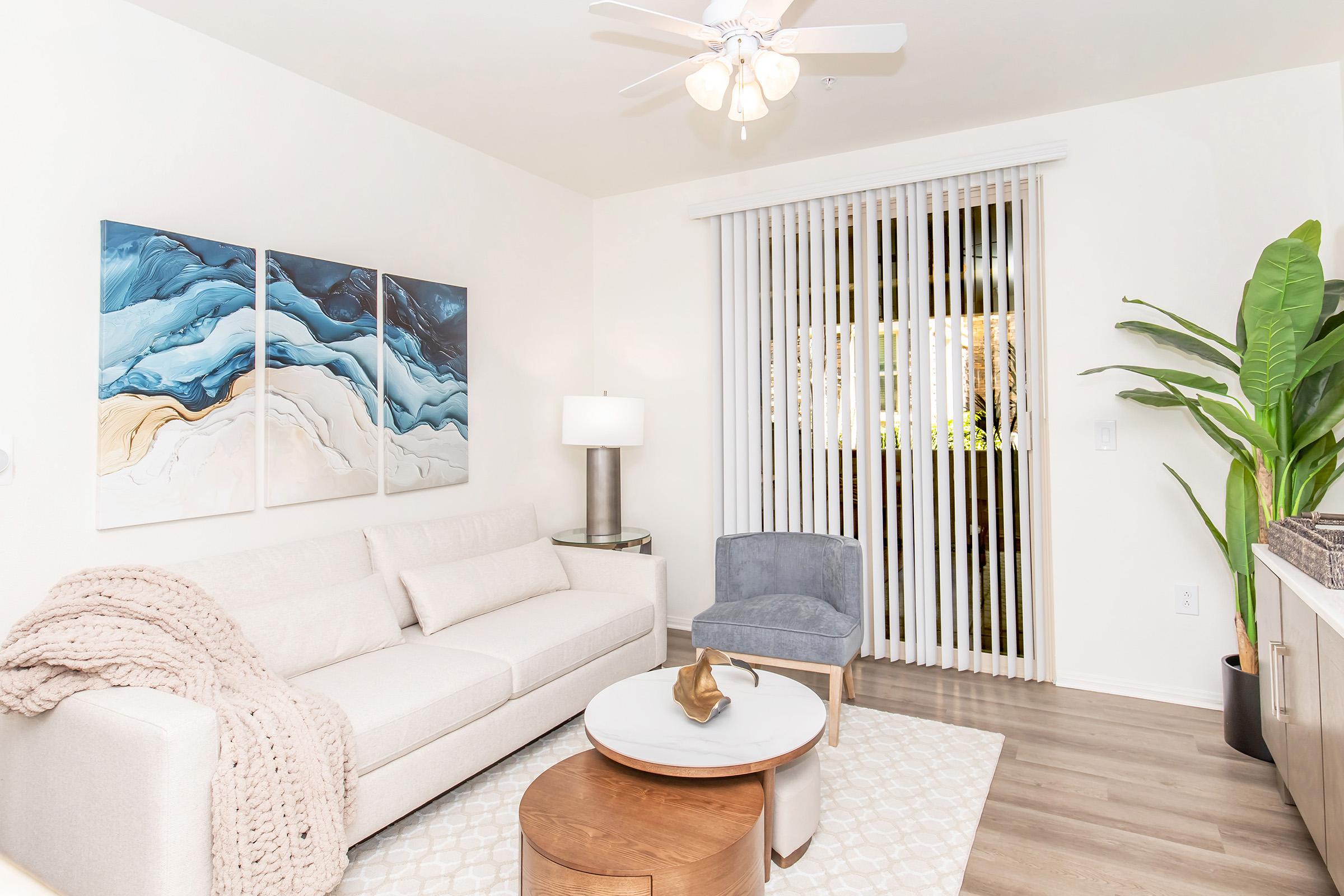A cozy living room featuring a light sofa with a decorative throw, a round coffee table, and a blue accent chair. Abstract artwork in shades of blue and beige adorns the wall. Natural light streams in through vertical blinds, and a potted plant adds a touch of greenery to the space.