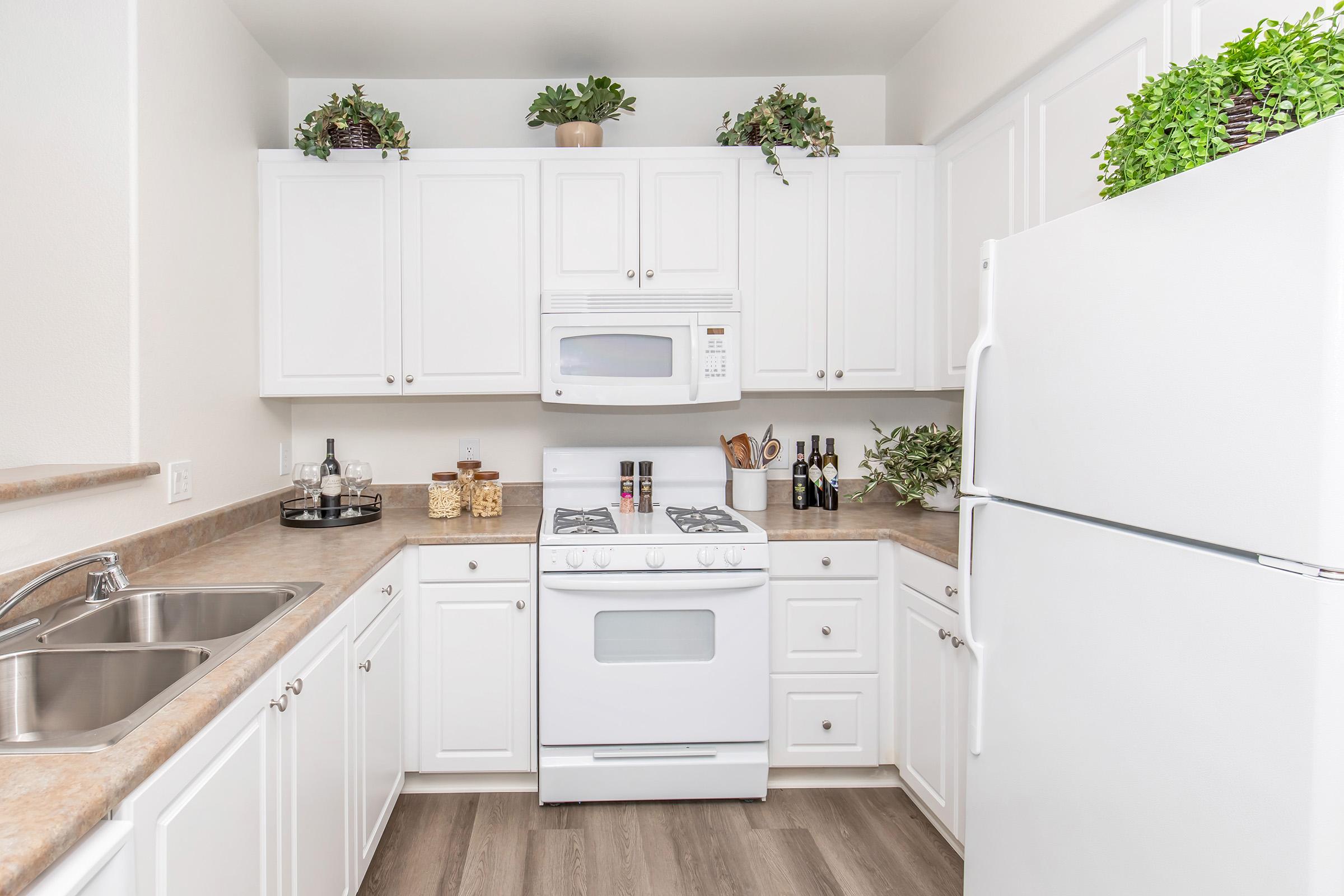 A modern kitchen featuring white cabinets and appliances, a white gas stove, and a refrigerator. The countertops are light-colored, adorned with decorative plants and kitchen essentials. There are jars of ingredients and wine bottles on display, creating a tidy and inviting cooking space with wood-like flooring.