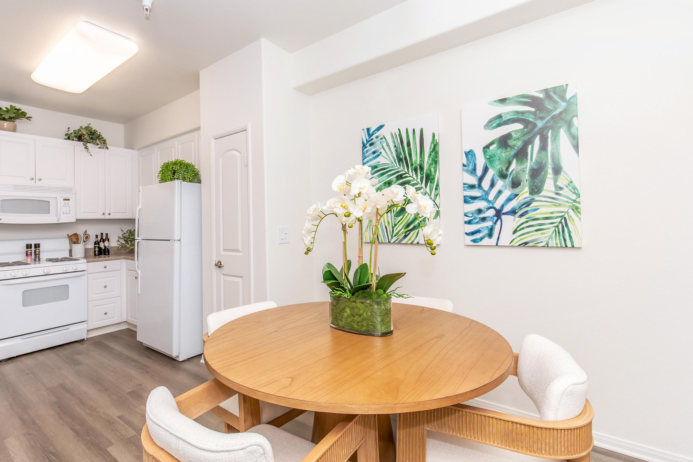 A modern kitchen area featuring a round wooden dining table with four white upholstered chairs. In the center, there is a vase of white orchids. The walls are adorned with two vibrant tropical leaf paintings, and a white refrigerator and cabinets can be seen in the background, along with decorative plants.