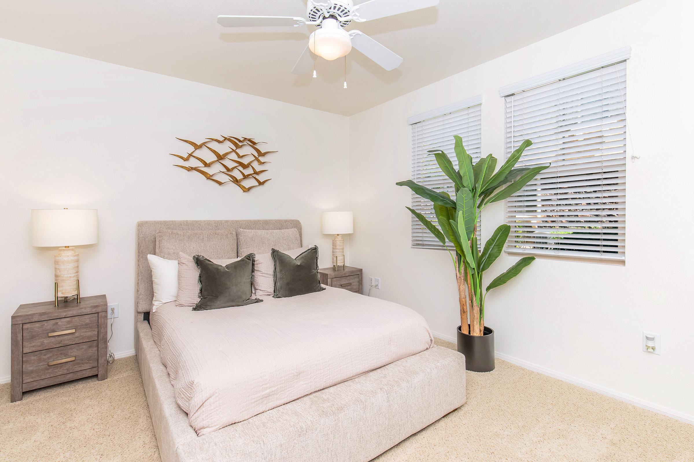 A cozy bedroom featuring a queen-sized bed with light bedding and decorative pillows, flanked by two modern nightstands with lamps. A stylish wall decoration made of metal fish hangs above the bed. The room has large windows with blinds, allowing natural light, and a tall potted plant in the corner adds a touch of greenery.