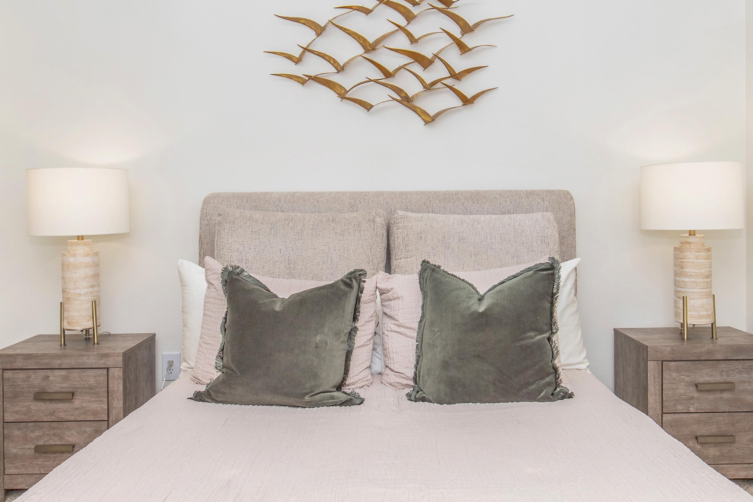 A cozy bedroom featuring a light-colored bed with plush pillows, two bedside tables with elegant lamps, and a decorative wall art piece resembling a flock of birds. The color palette is neutral, creating a serene and inviting atmosphere.