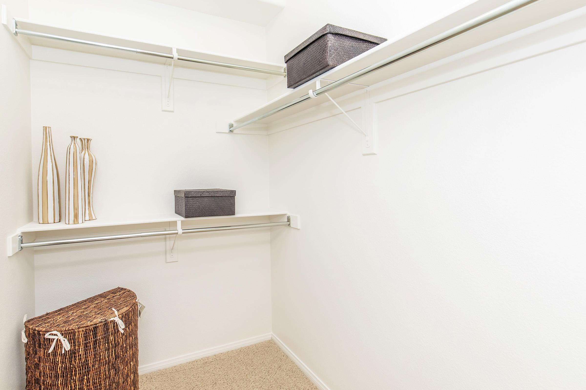 A clean and organized closet featuring two empty shelves, a woven basket, and decorative vases. The walls are painted white, and the floor is carpeted. One shelf holds a small storage box while the other shelf remains clear.