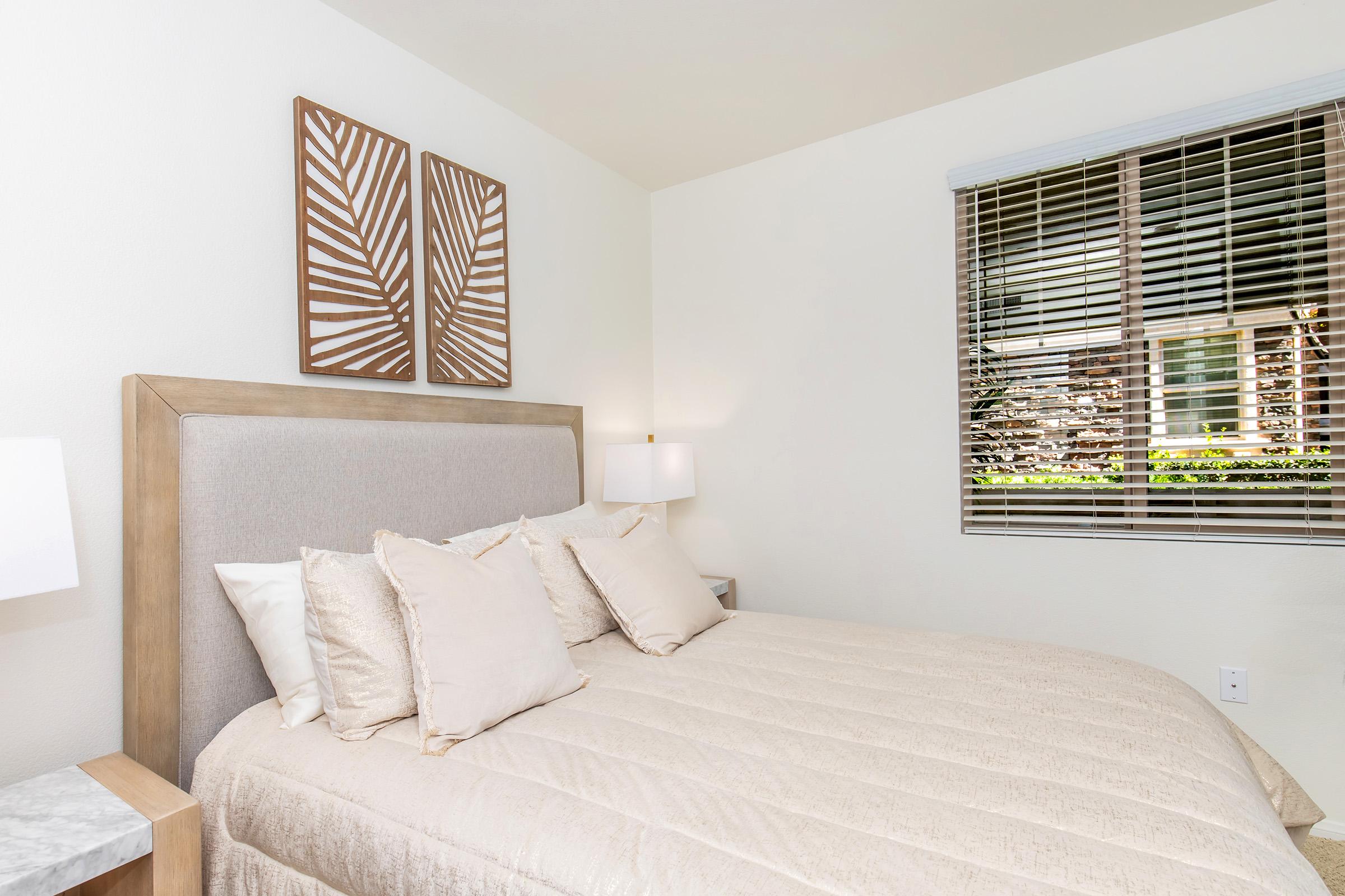 A cozy bedroom featuring a beige bedspread and multiple decorative pillows on a bed with a soft headboard. There are two framed palm leaf art pieces on the wall above the bed, and a window with blinds providing natural light. A nightstand with a lamp is positioned next to the bed, creating a tranquil atmosphere.