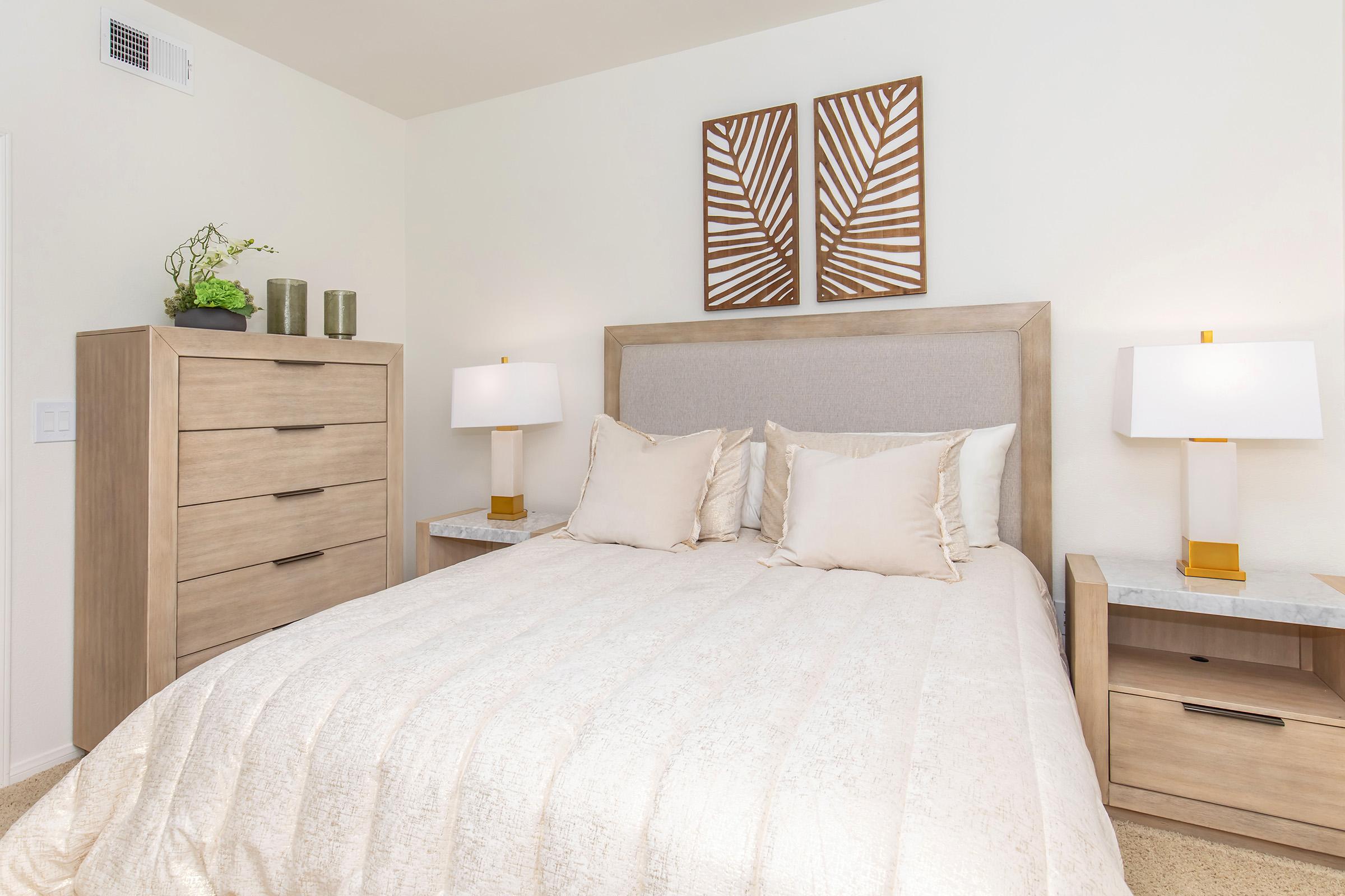 A cozy bedroom featuring a queen-sized bed with light-colored bedding, two decorative pillows, and a headboard. There are matching bedside tables with lamps, a wooden dresser, and wall art depicting leaf designs. Natural light illuminates the room, enhancing its warm and inviting atmosphere.