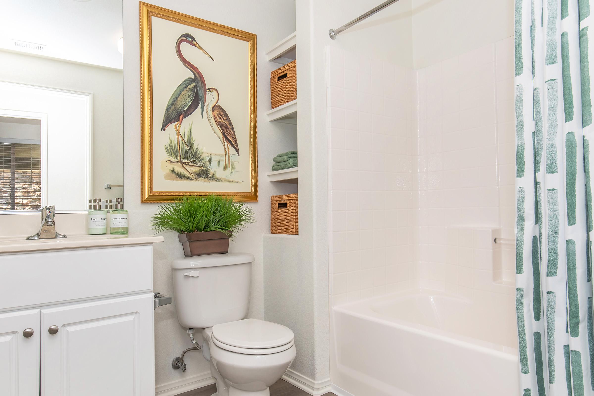 A modern bathroom featuring a white bathtub and toilet, a large decorative print of two herons on the wall, and shelves displaying neatly folded towels and woven baskets. The shower curtain has a green and white pattern, and there's a small potted plant adding a touch of greenery to the space.