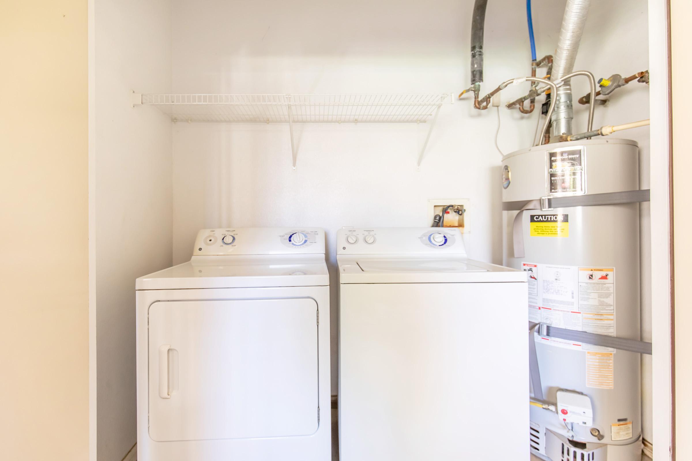 A laundry room featuring a stacked washer and dryer on the left, and a water heater on the right. The space includes a wire shelf above the appliances for storage. The walls are painted a light color, creating a clean and functional atmosphere.