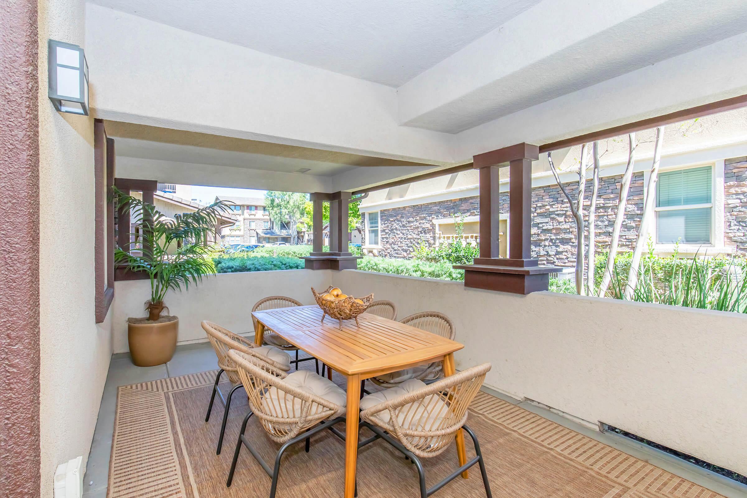 A covered patio area featuring a wooden dining table surrounded by wicker chairs. A potted plant adds greenery, while stone wall accents and large windows create an inviting atmosphere. Natural light filters in, enhancing the outdoor dining experience.
