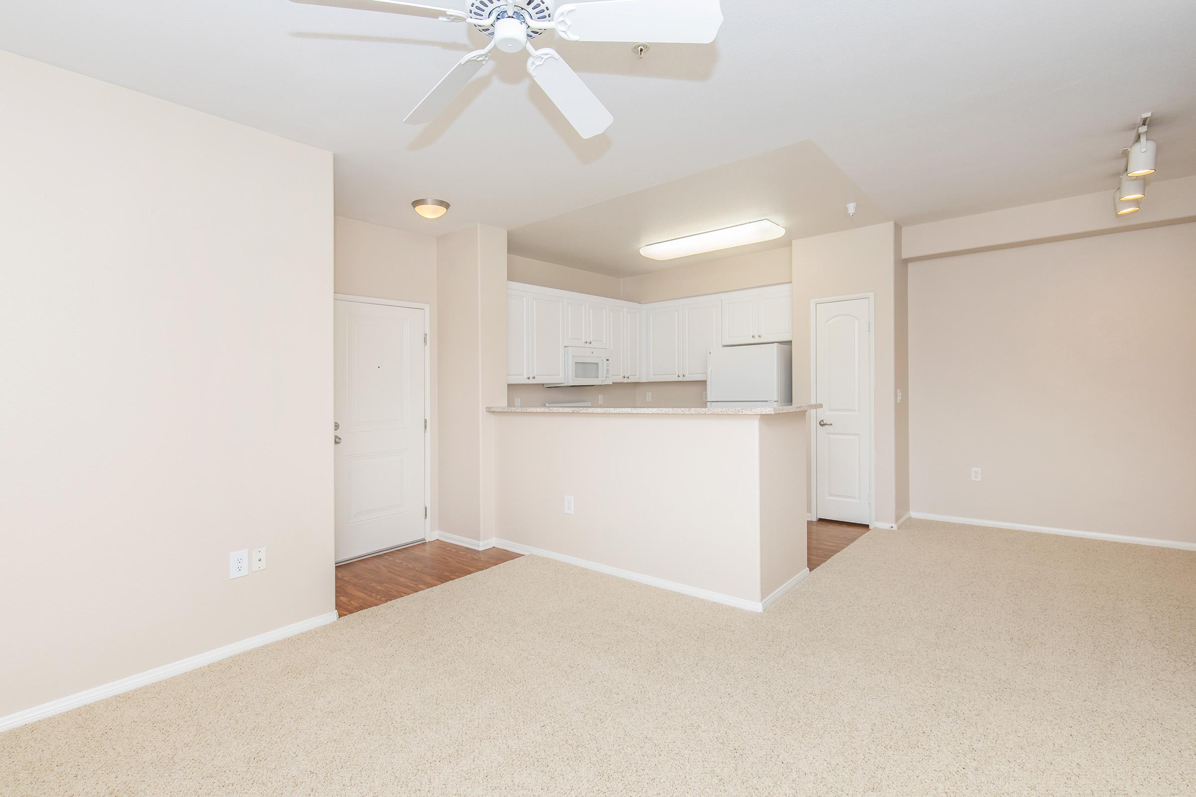 A spacious, neutral-colored living area with beige carpet and walls. There is a light ceiling fan, a white kitchen with wooden flooring visible in the background, and a doorway leading to another room. The area is well-lit with natural light and features simple, modern design elements.