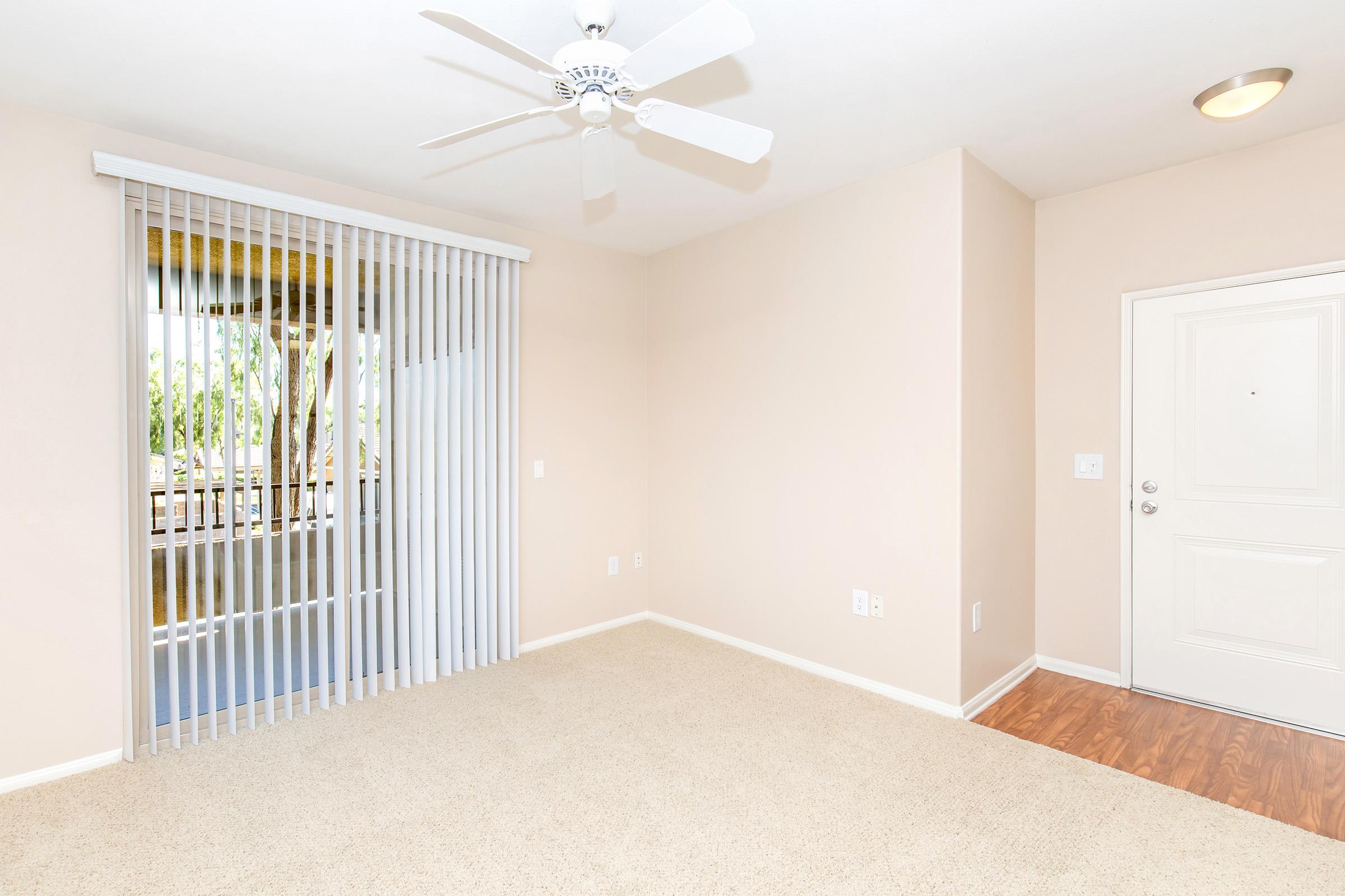 Empty room with beige walls and carpet, featuring a ceiling fan and vertical blinds. A door leads to an outdoor area, and there is a light fixture on the ceiling. The space feels bright and inviting, with natural light streaming in.