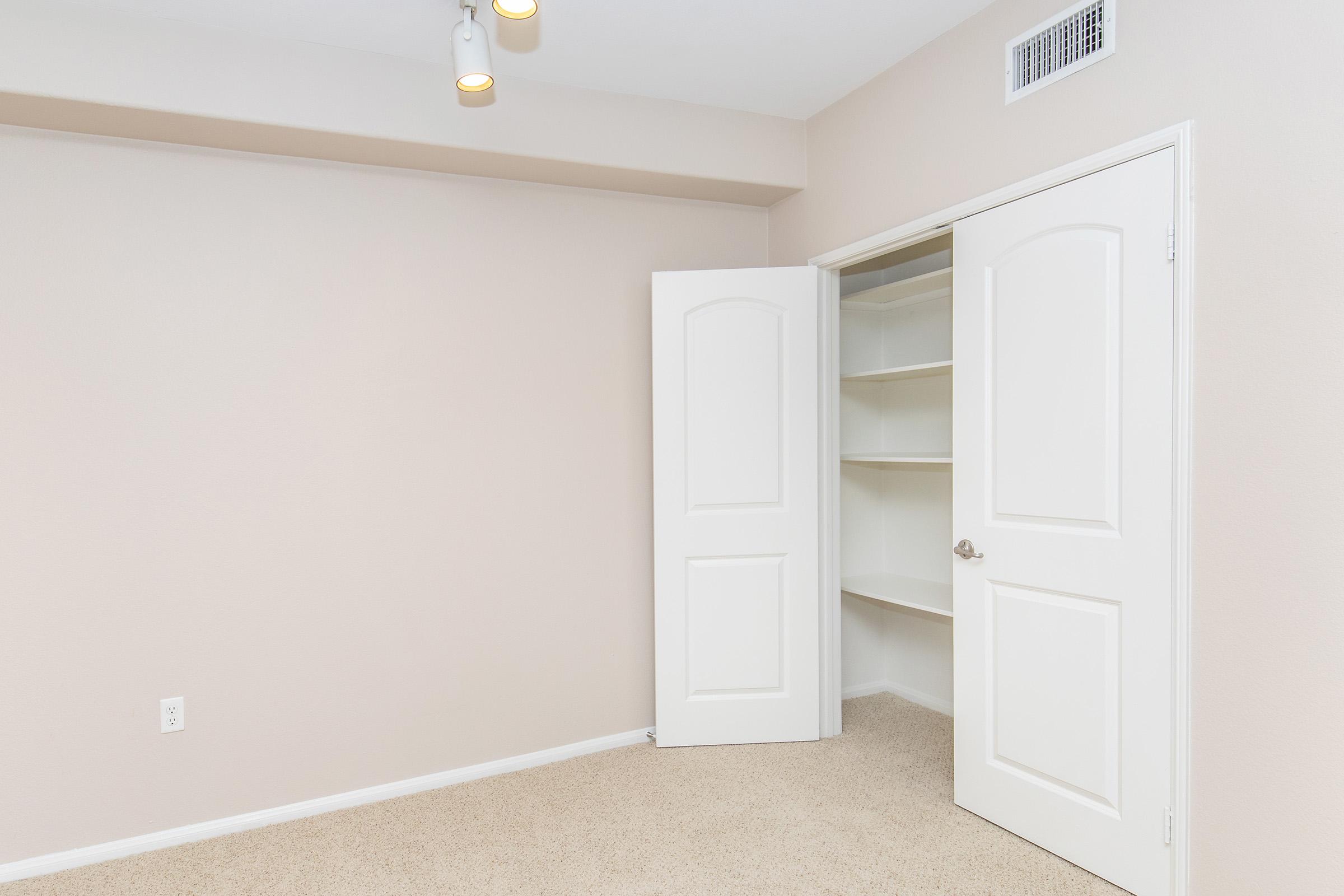 A well-lit empty room with beige walls and carpet, featuring an open closet with shelves. Two ceiling lights illuminate the space, and a single light switch is visible on the left wall. The room has a simple design and no furniture, creating a clean and minimalistic appearance.