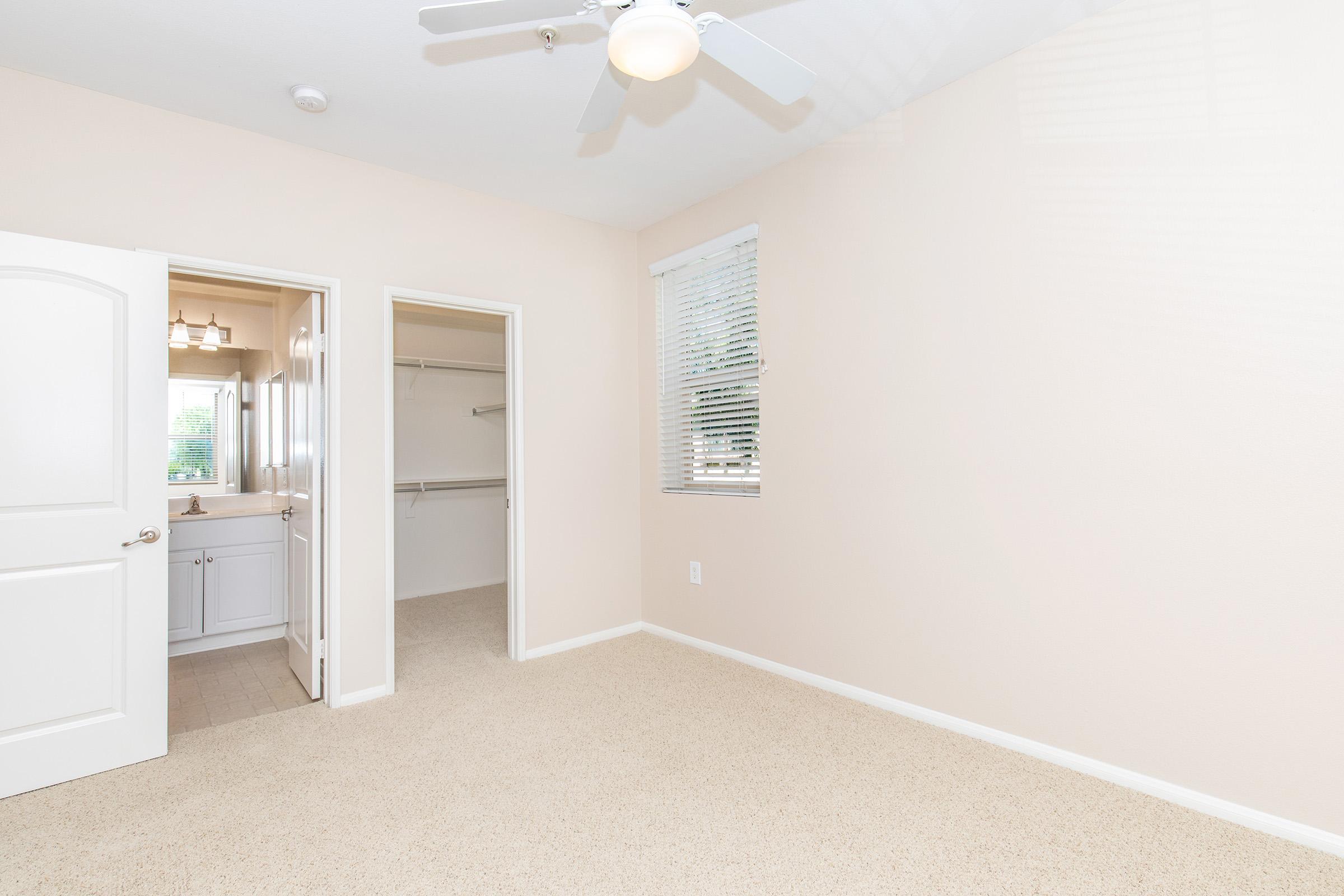 A bright and airy bedroom featuring beige walls, a ceiling fan, and carpeted floor. To the left, there is a doorway leading to a bathroom with a sink and a closet space. A window allows natural light to fill the room, enhancing the spacious feel.