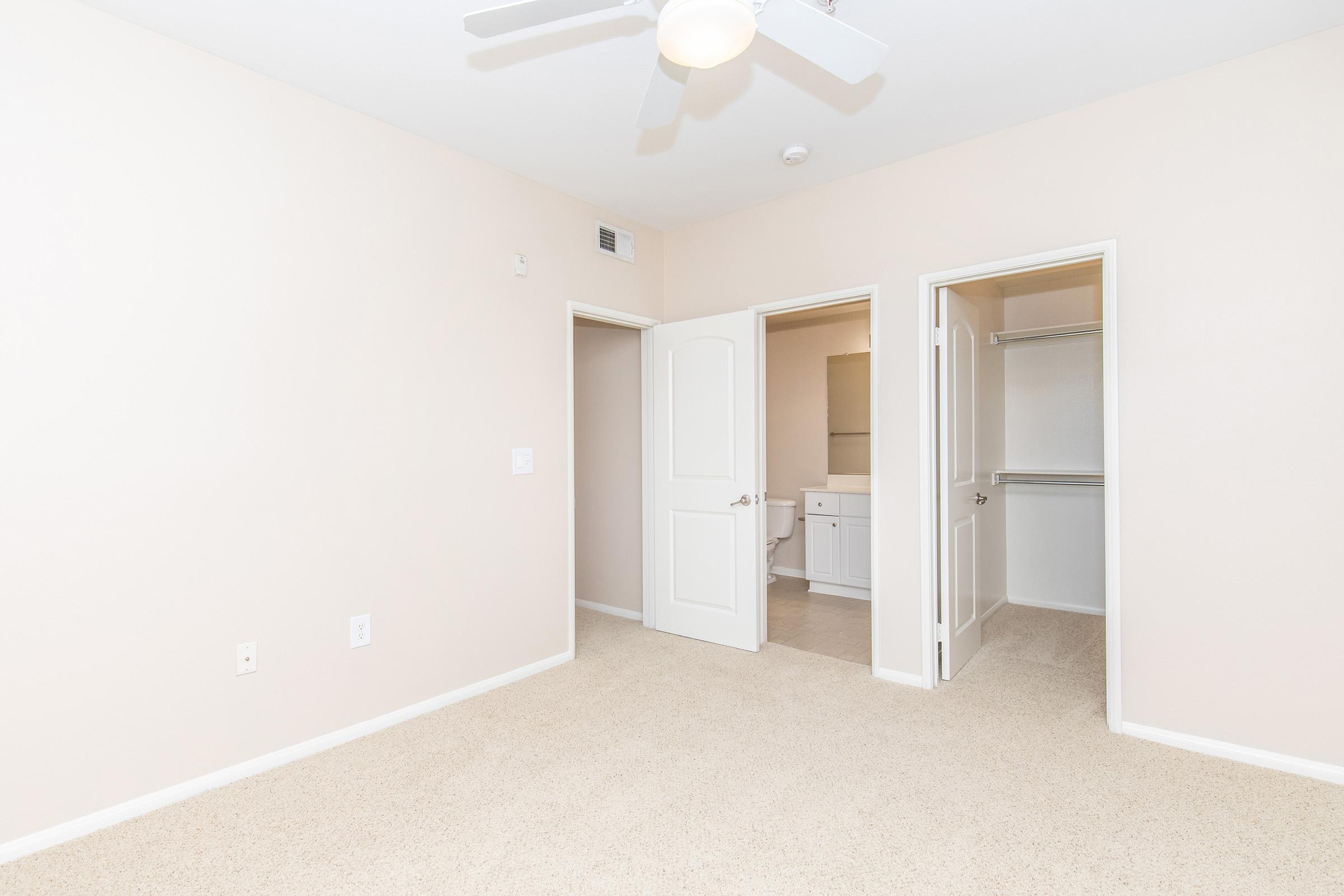 A light-colored room featuring beige walls and carpet, with a ceiling fan. There are two open doors leading to a bathroom and closet, providing a spacious feel. The room is well-lit and minimalist, suitable for various uses.