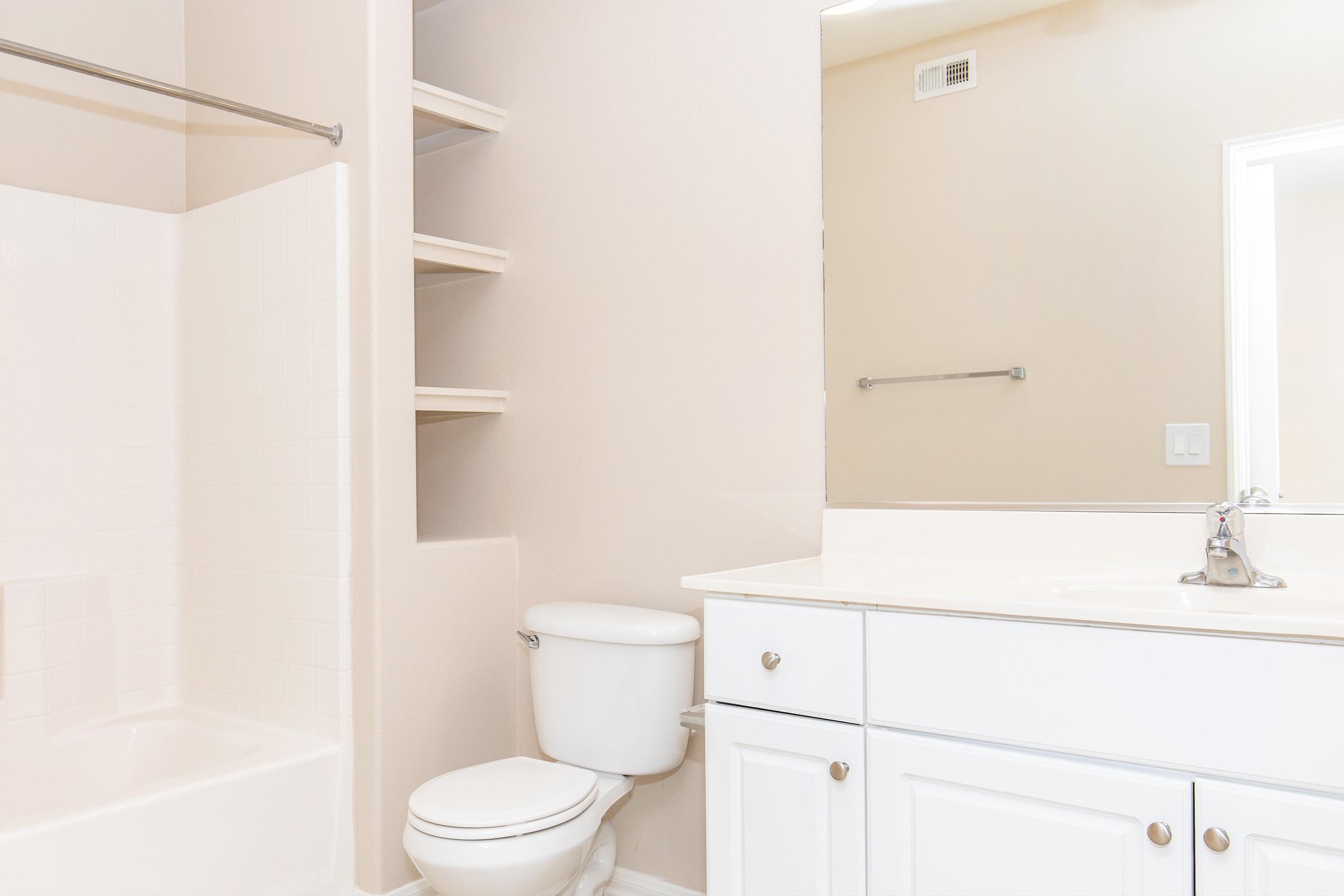 A clean, modern bathroom featuring a white bathtub and shower, a white toilet, and a vanity with a sink. There are light-colored walls and a large mirror above the sink. Shelving is visible on the left side, providing storage space. The overall design is simple and functional.