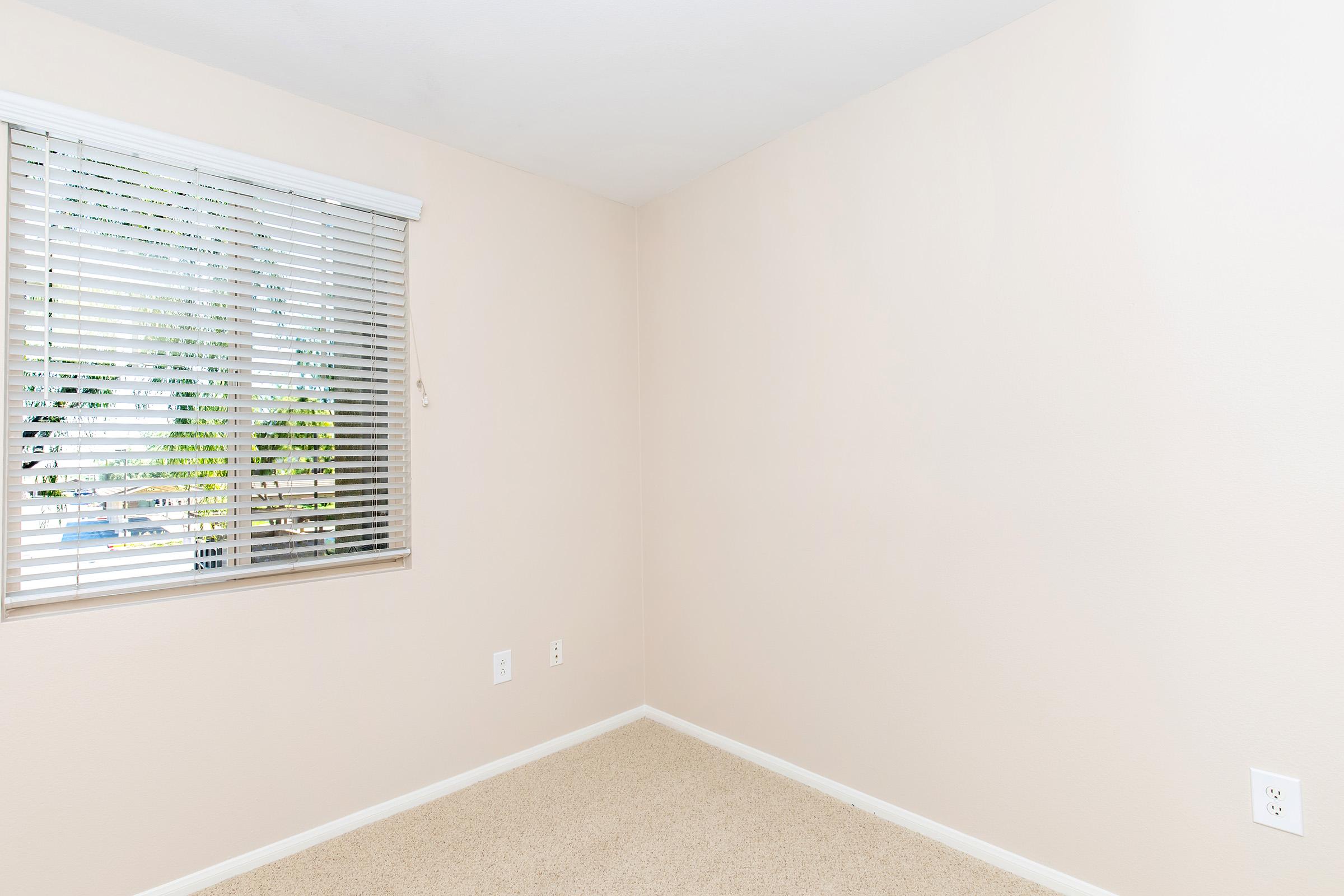 An empty room with a light beige wall and beige carpet. A window with white blinds allows natural light to enter, showing partly visible greenery outside. The room is unfurnished, creating a spacious and neutral atmosphere, ideal for various uses or decoration.