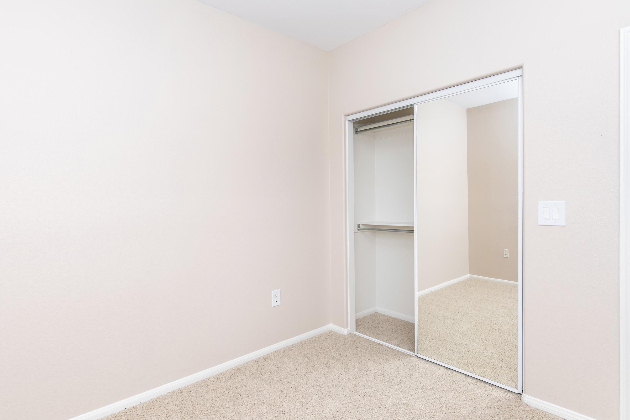 Empty room with light beige walls and a mirrored closet. The floor is covered with beige carpet, and there are no furnishings or decorations. The closet features sliding mirrors, creating a spacious look in the simple, uncluttered space.