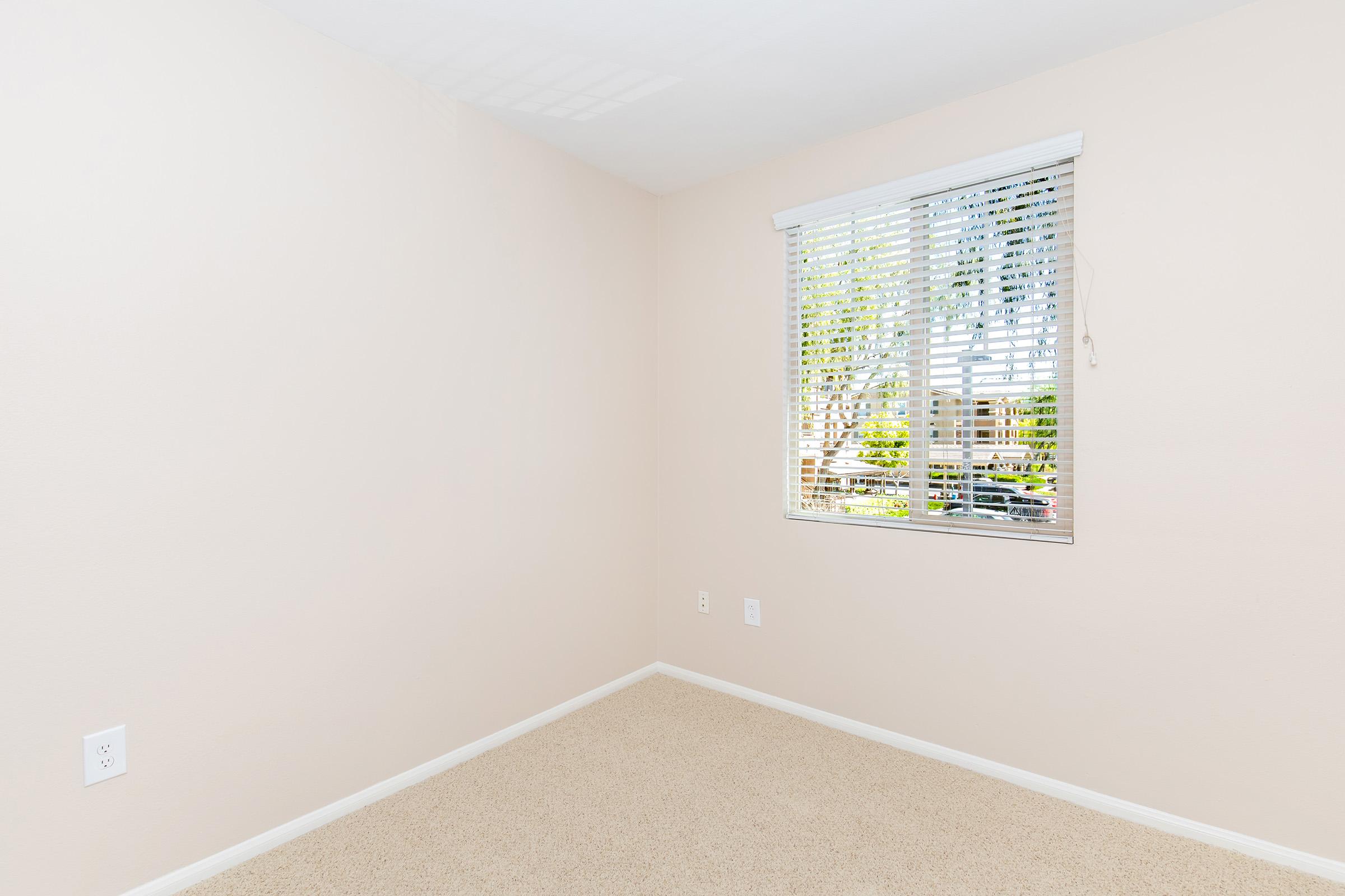 Empty room with light beige walls and neutral carpet. A window with white blinds allows natural light to enter, revealing greenery outside. The space is uncluttered, ideal for a living area or bedroom. Quiet and bright, it offers a blank canvas for decorating.