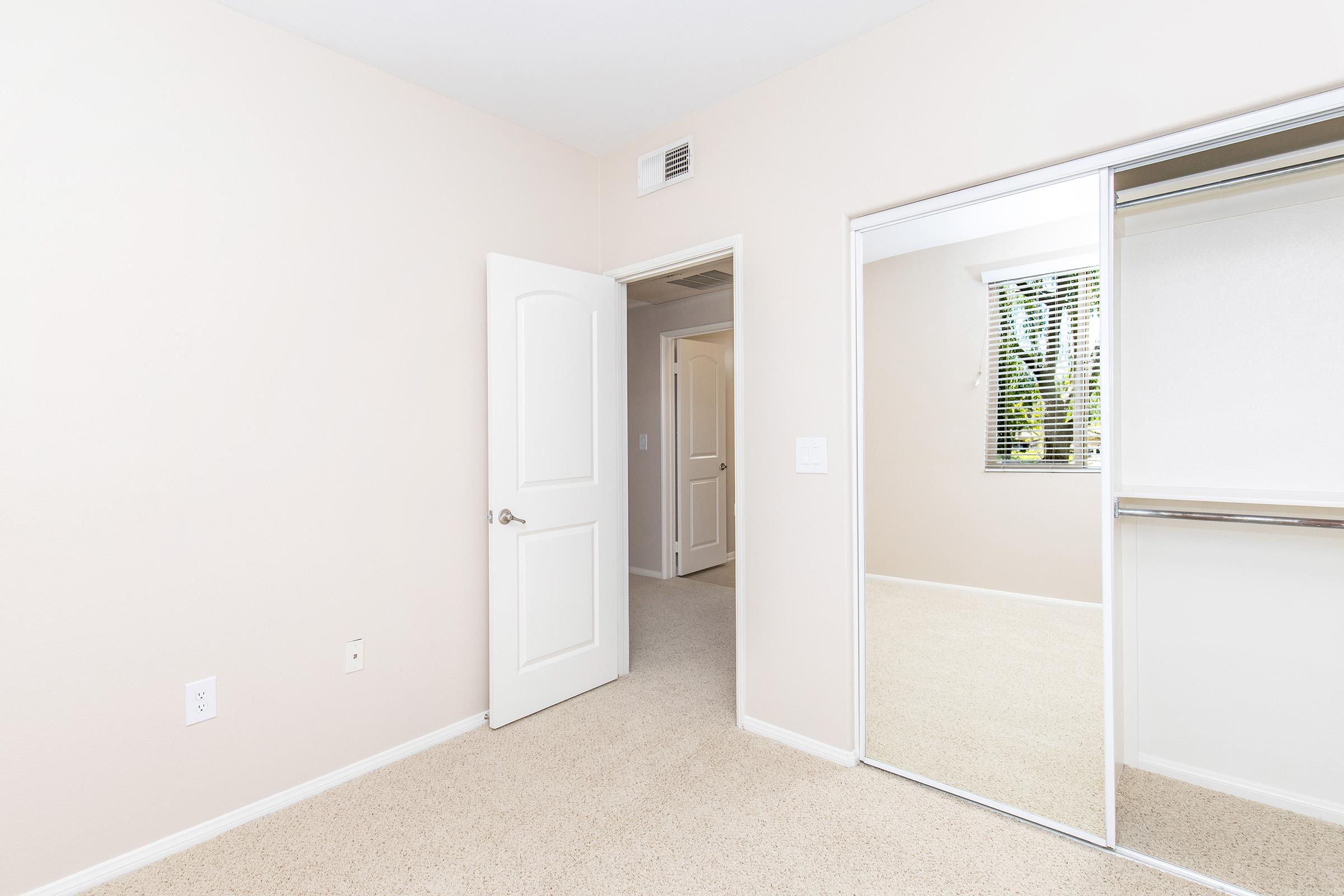 A light-colored room with beige walls and carpet. The room features a doorway leading to another space, a large mirrored closet, and a window with blinds allowing natural light. The overall atmosphere is bright and airy, suitable for various arrangements and uses.