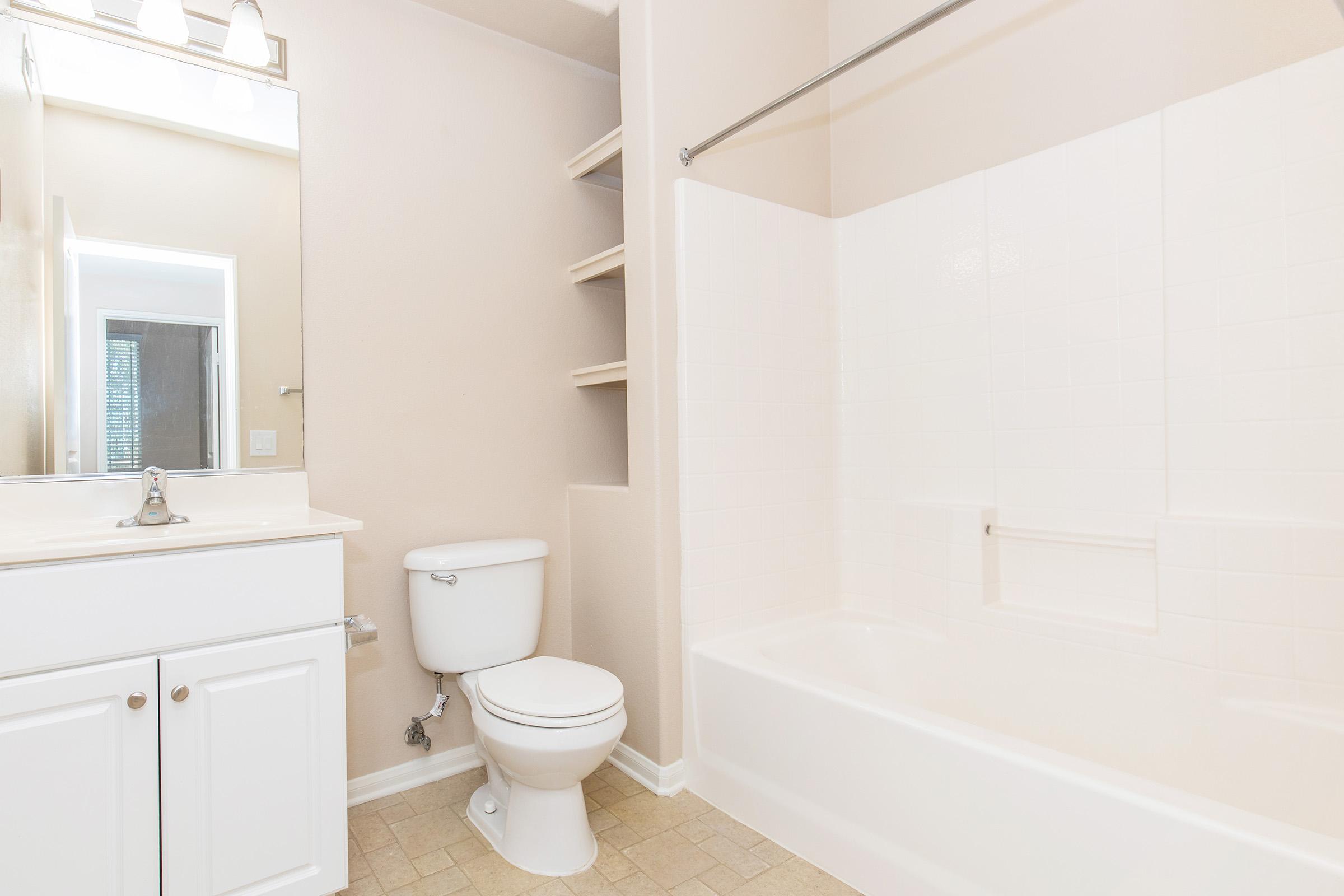 Bright, clean bathroom featuring a white bathtub and shower, a vanity with a sink, a toilet, and a large mirror. Light beige walls complement the neutral tile flooring. There are shelves for storage next to the bathtub, enhancing the functionality of the space.
