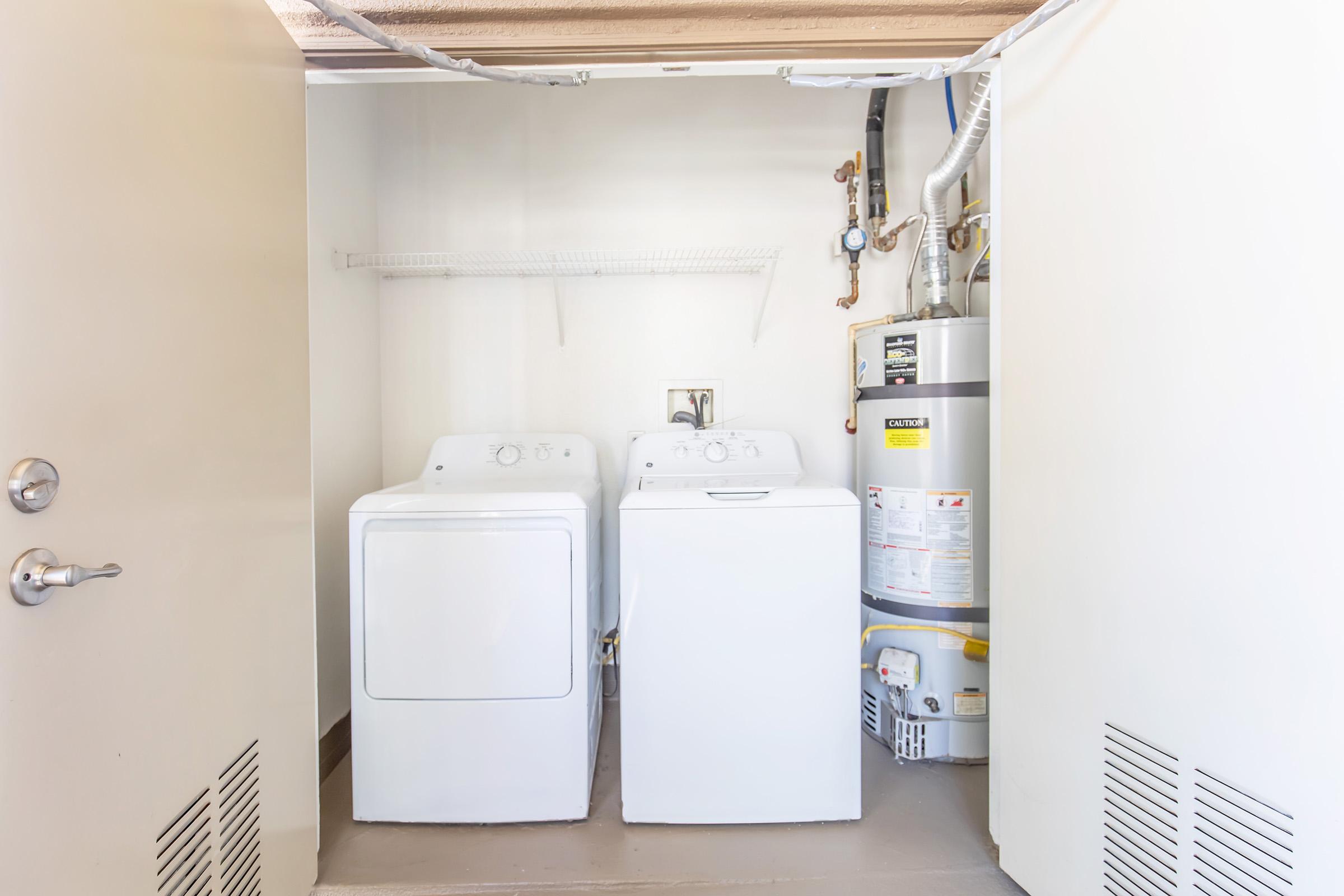 A clean laundry room featuring a white washer and dryer side by side. There is a water heater on the right and a shelf above the appliances. The walls are painted light colors, and there is a door leading out of the space. The floor is simple and uncluttered, creating an organized appearance.